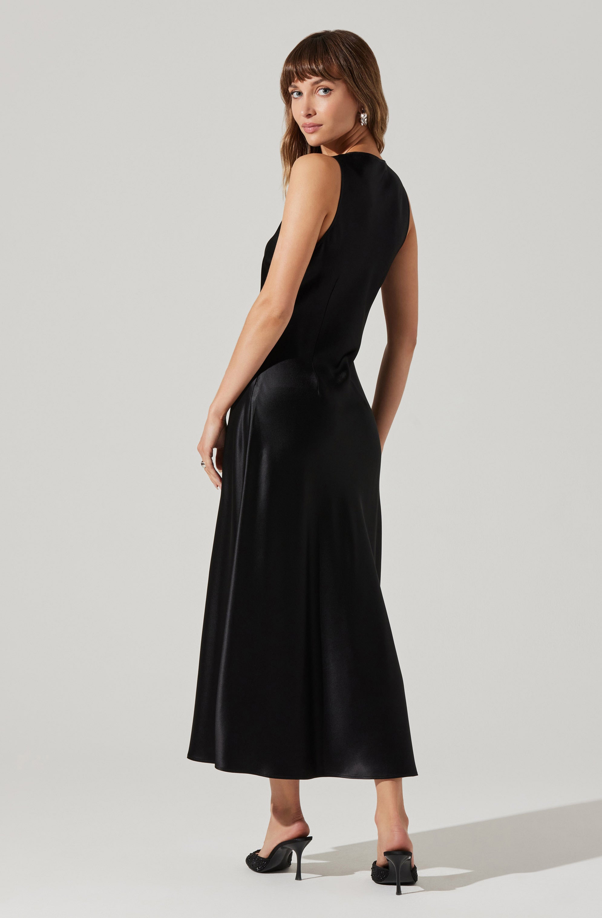 A woman is wearing a sleeveless, floor-length black satin gown with a fitted bodice and a slightly flared skirt. She has paired the dress with black high-heeled shoes that have an open toe and a bow detail at the front. She is accessorized with large, dangling earrings.