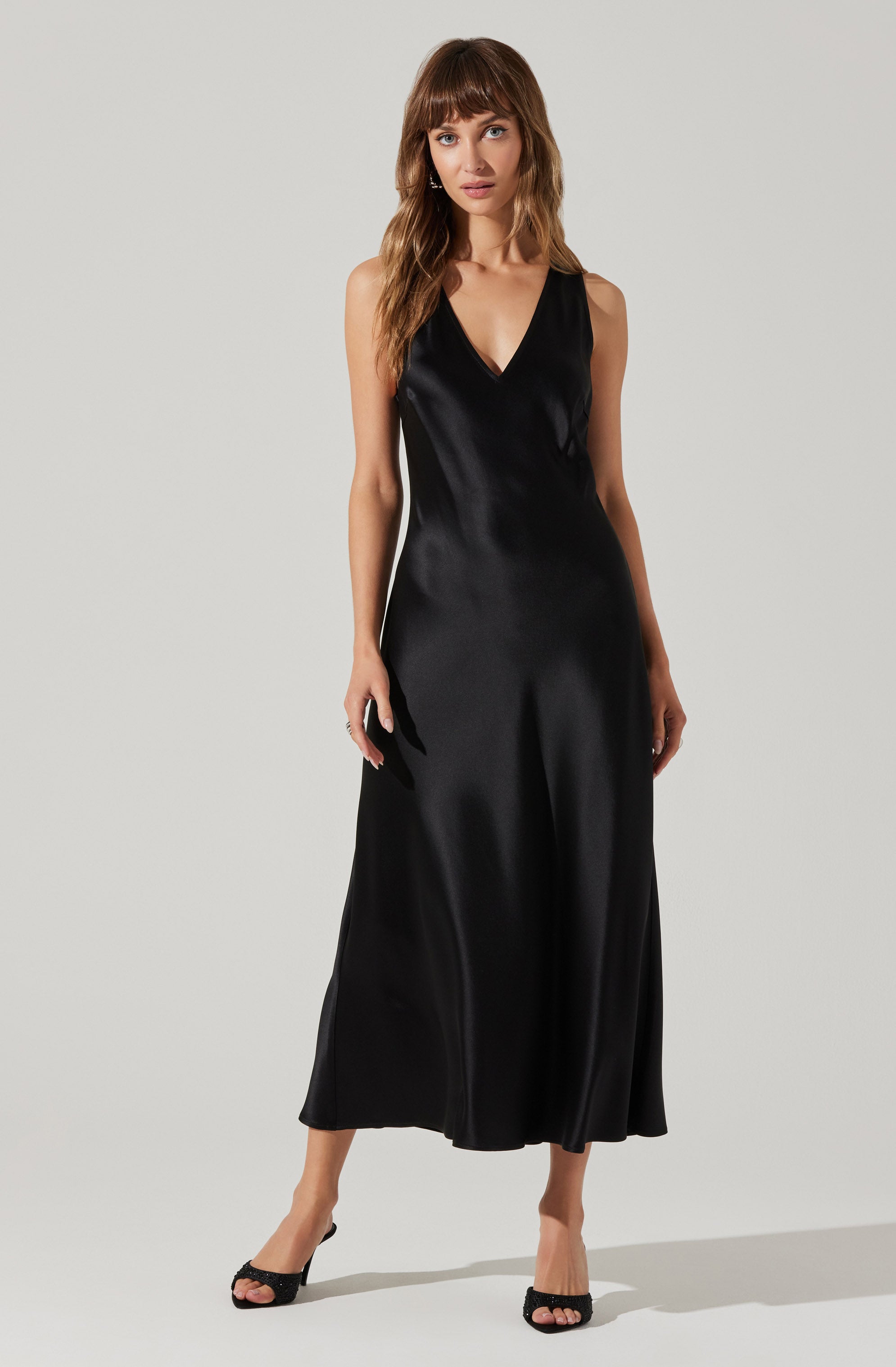 A woman is wearing a sleeveless, black satin maxi dress with a V-neckline. She is also wearing black high-heeled sandals.
