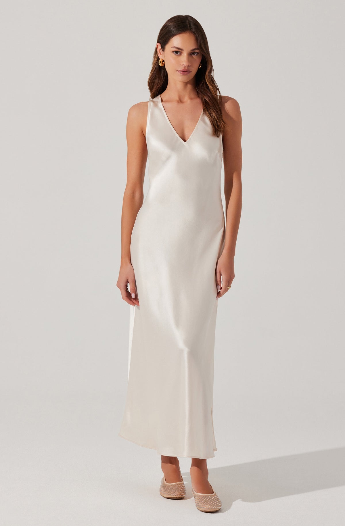 A woman is wearing a sleeveless, full-length, cream-colored satin dress with a V-neckline. She has on gold hoop earrings and nude-colored woven heels.