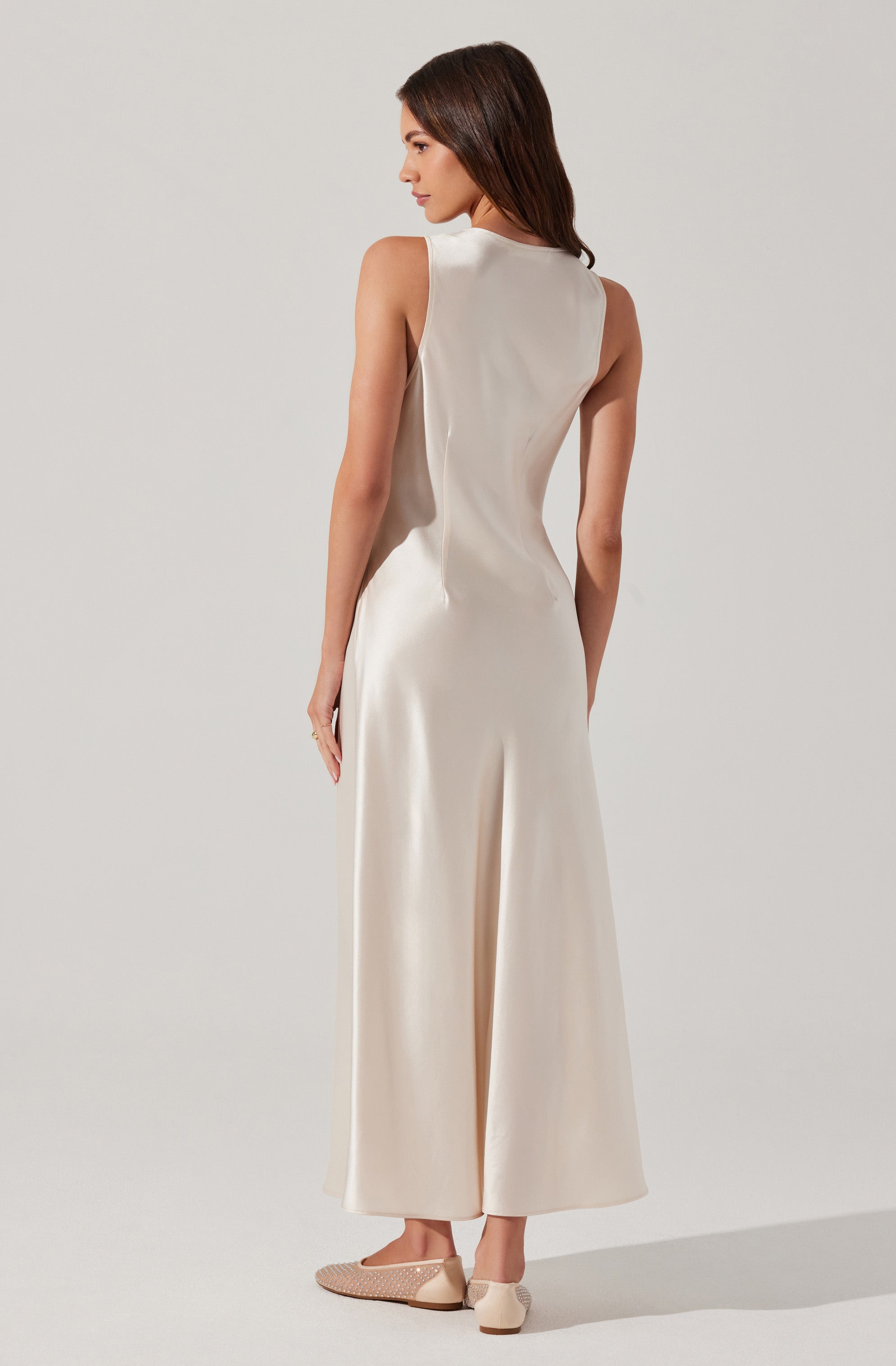 A woman is wearing a sleeveless, high-neck, floor-length cream-colored dress made of a smooth, shiny fabric. She is also wearing beige flats with a textured pattern.