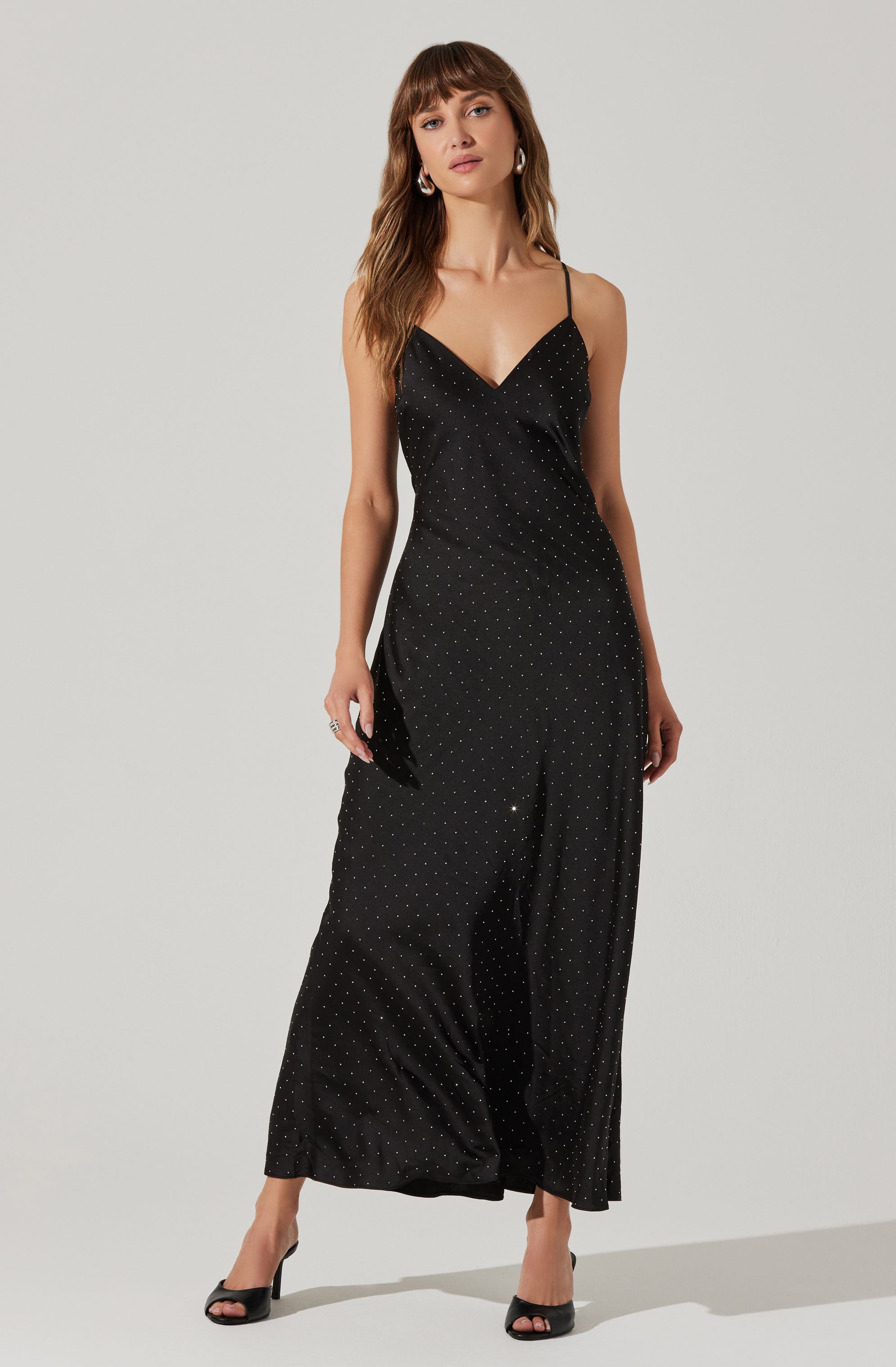 A woman is wearing a black, floor-length dress with thin spaghetti straps. The dress features a subtle pattern of small, evenly spaced, light-colored dots. The fabric appears silky and smooth, giving it an elegant and dressy appearance. She is also wearing black open-toe high heels.