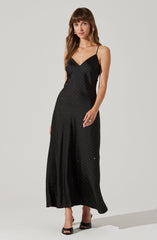 A woman is wearing a black, floor-length dress with thin spaghetti straps. The dress features a subtle pattern of small, sparkling dots throughout. She is also wearing black high-heeled shoes.