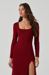 A woman is wearing a long, fitted, ribbed dress in a deep red color. The dress features a scoop neckline, long sleeves, and a high slit on the left side that reveals her leg.