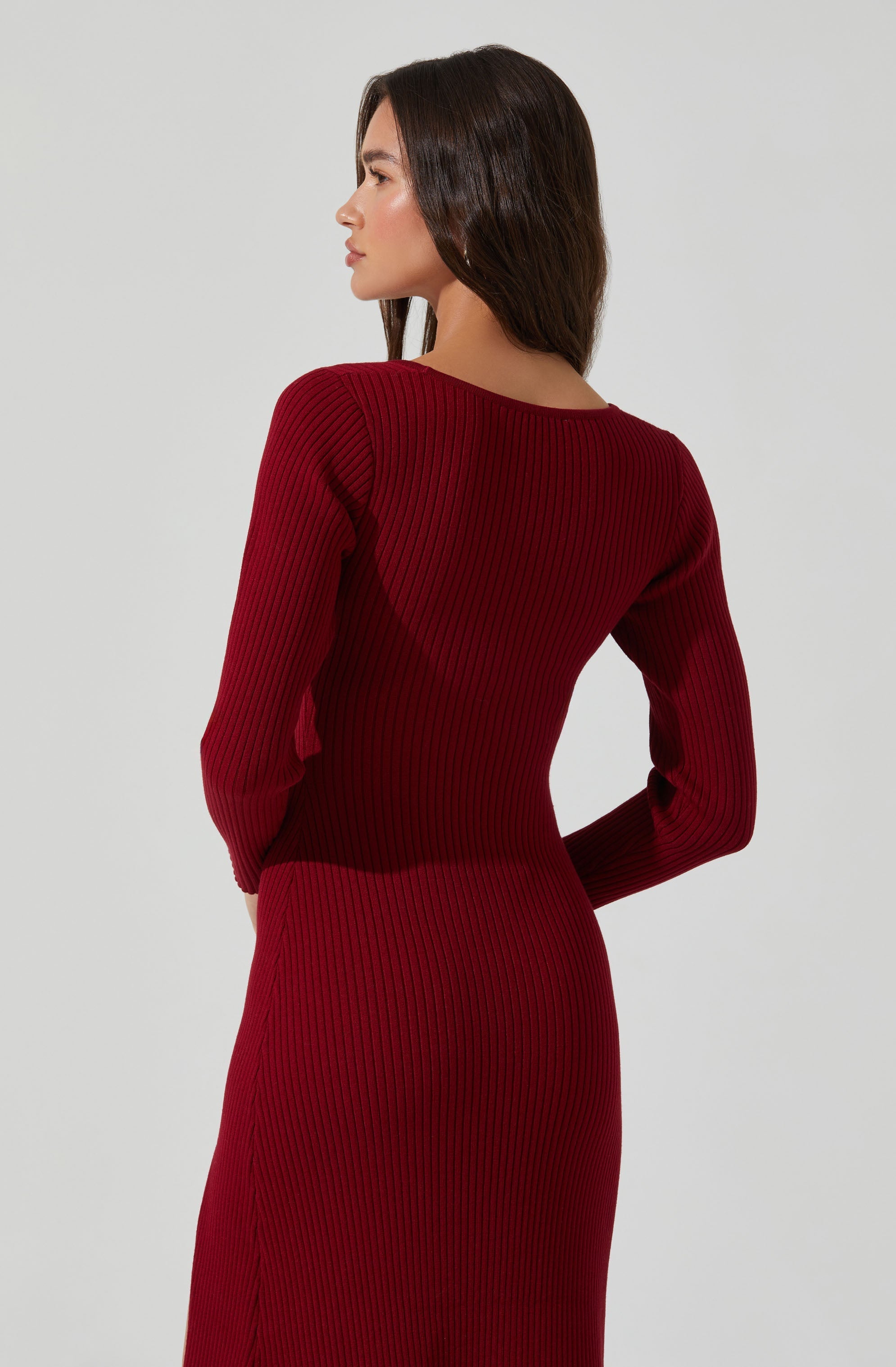A woman is wearing a fitted, long-sleeved, ribbed dress in a deep red color. The dress has a round neckline and features vertical ribbing throughout.