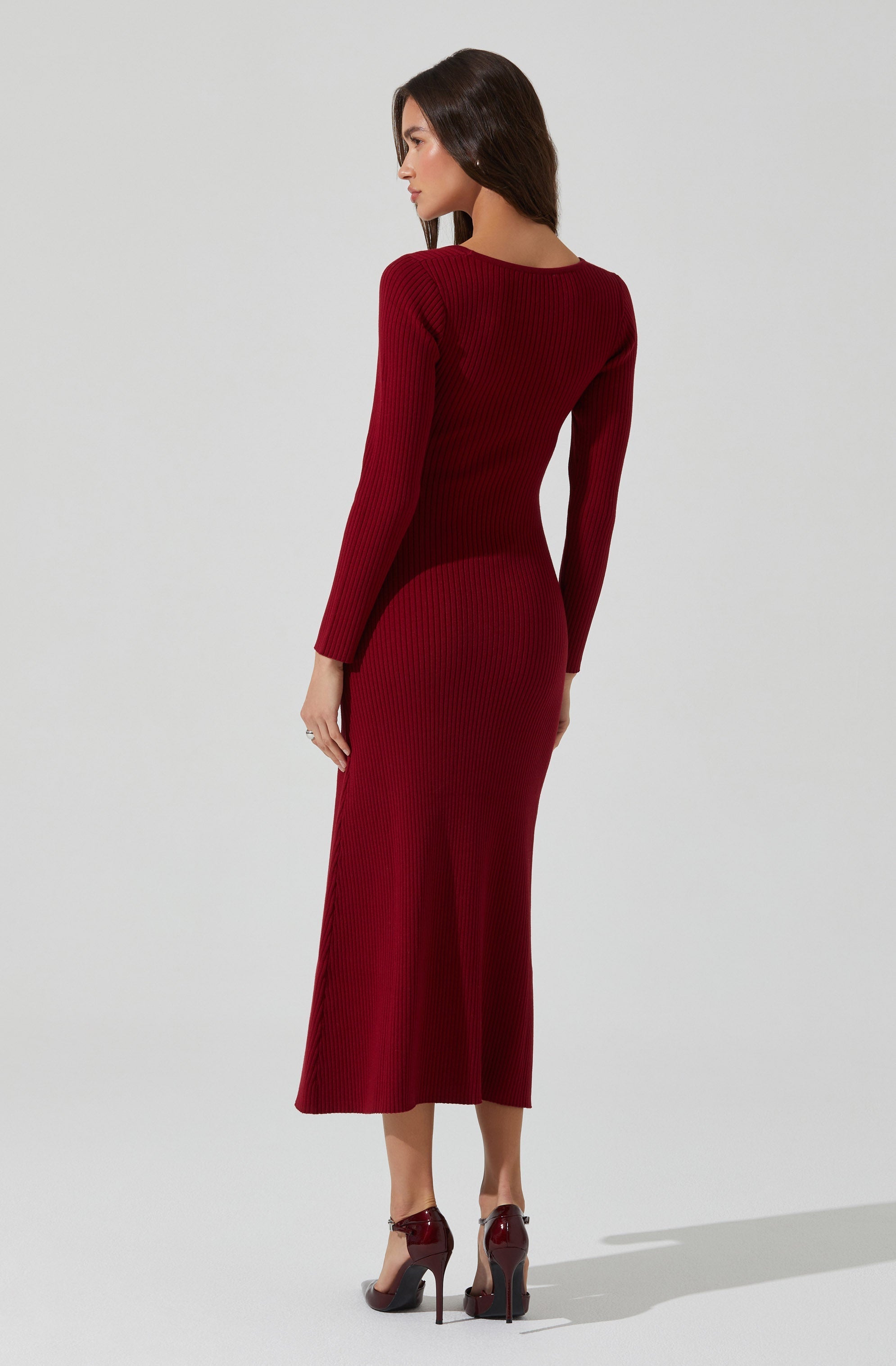 A woman is wearing a fitted, long-sleeved, ribbed knit dress in a deep red color that extends to her ankles. She is also wearing matching high-heeled shoes in a similar shade of red.