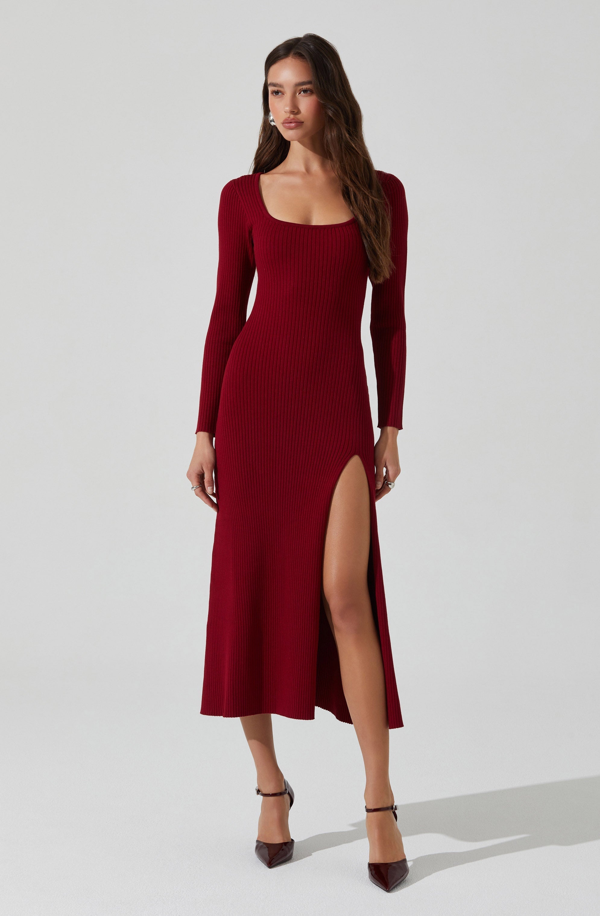 A woman is wearing a long, fitted, ribbed knit dress in a deep red color. The dress features long sleeves, a square neckline, and a high slit on the left side, revealing her leg. She is wearing dark-colored high-heeled shoes with ankle straps.