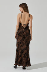 A woman is wearing a long, form-fitting dress with a leopard print pattern in shades of brown and black. The dress has a backless design with thin spaghetti straps and a lace-up detail at the waist. She is also wearing black high-heeled shoes.