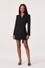 A woman is wearing a black blazer-style dress with a tailored fit, featuring a notched lapel collar and long sleeves. The dress has a unique design with a tied belt on the right side, creating pleats that fall asymmetrically. She is also wearing pointed high-heeled shoes with a black and white pattern.