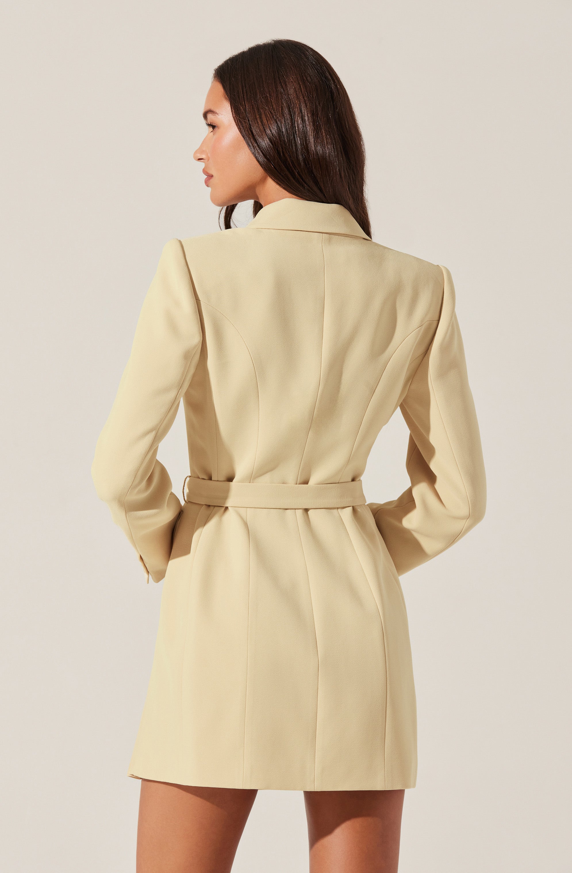 A woman is wearing a beige tailored blazer dress with a belt at the waist. The blazer features structured shoulders and vertical seam detailing on the back.