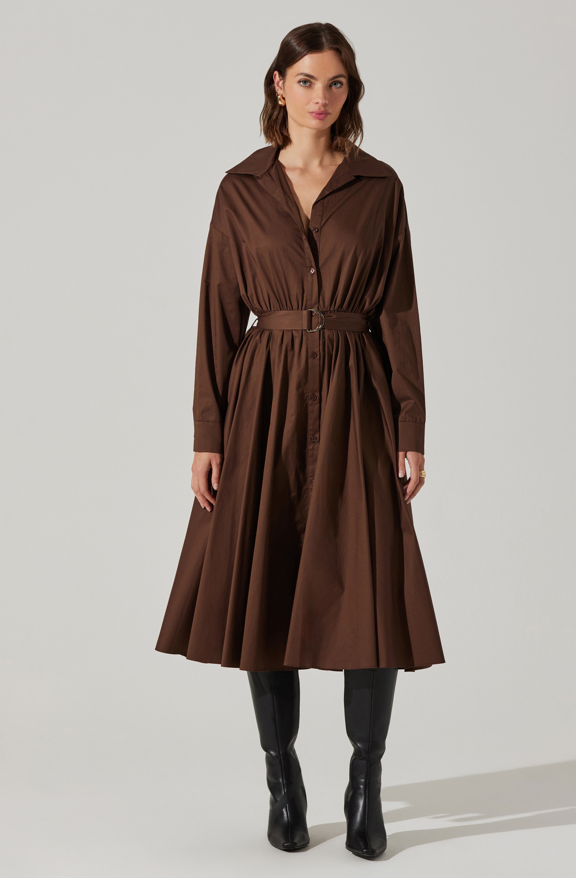 A woman is wearing a long brown shirt dress with a collar, button-down front, and long sleeves with cuffs. The dress features a matching belt at the waist, creating a gathered effect, and the skirt has pleats that give it a full, flowing silhouette. She is pairing the dress with knee-high black boots.