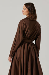 A woman is wearing a long, brown, shirt-style dress with a matching belt at the waist. The dress has long sleeves that extend to her wrists, with buttoned cuffs. The fabric appears to be smooth and slightly shiny.