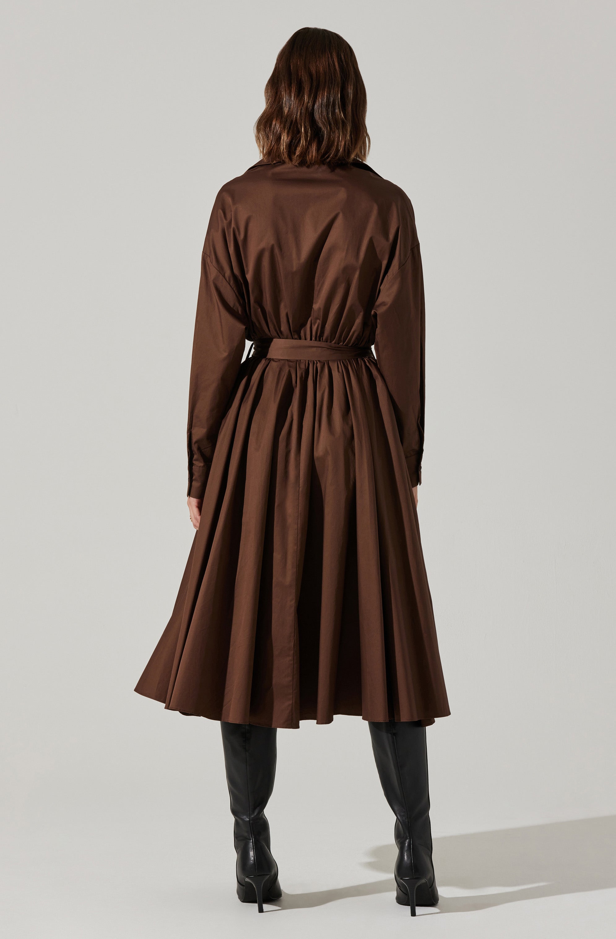 A woman is wearing a long, brown dress with a gathered waist and full skirt that extends to mid-calf. The dress has long sleeves and a collar, creating a structured, elegant look. She is also wearing tall black leather boots that reach just below the knee.