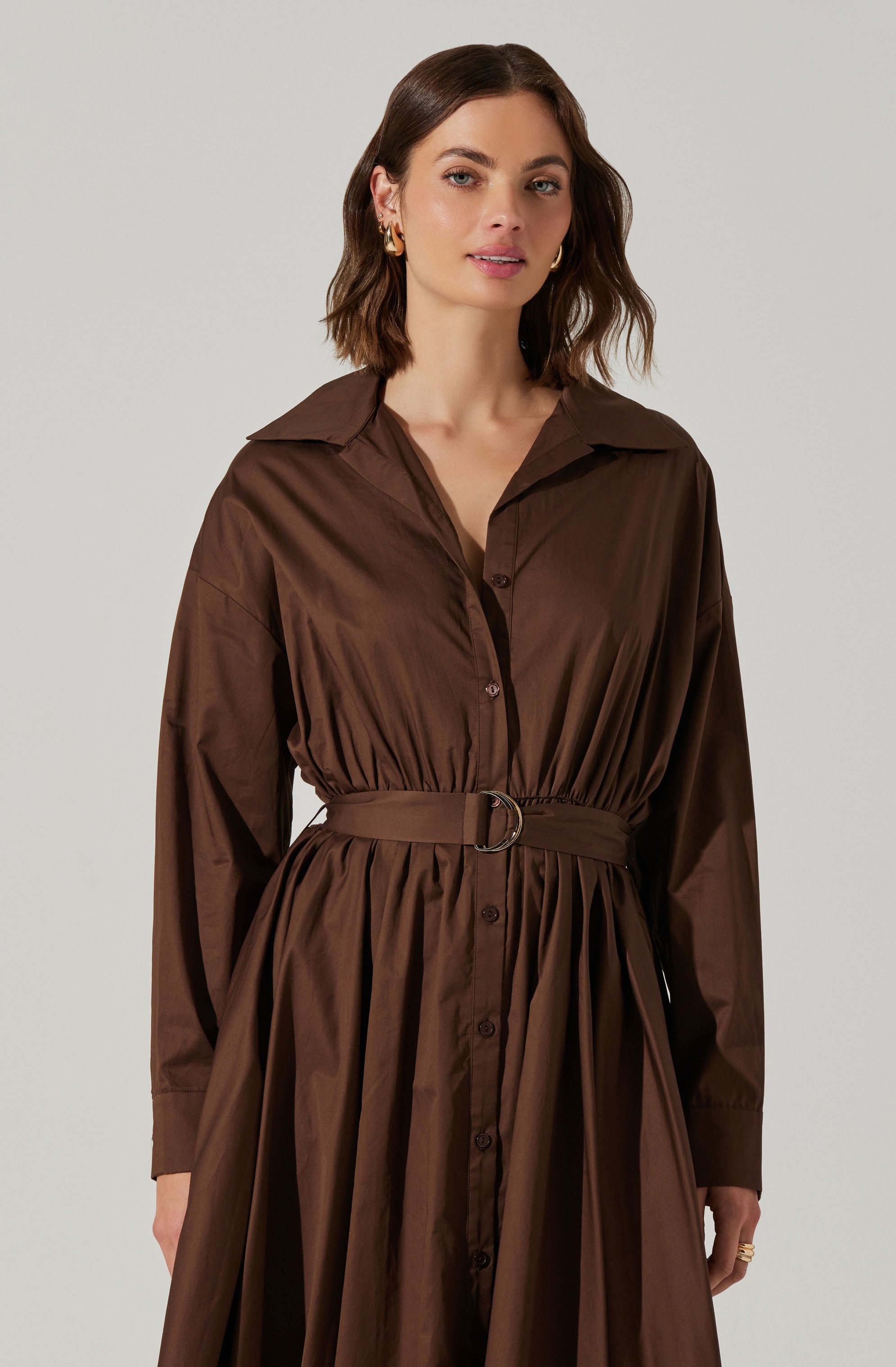 A woman is wearing a long, brown shirt dress made of shiny, lightweight fabric. The dress features a collar, button-down front, and long sleeves. It is cinched at the waist with a matching brown belt that has a silver D-ring buckle, creating a gathered effect around the waistline.