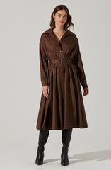 A woman is wearing a long, brown shirt dress with a collar and button-down front. The dress has long sleeves and is cinched at the waist with a matching belt that features a silver buckle. The skirt of the dress is voluminous and pleated, extending to a midi length. She is also wearing black, knee-high leather boots.