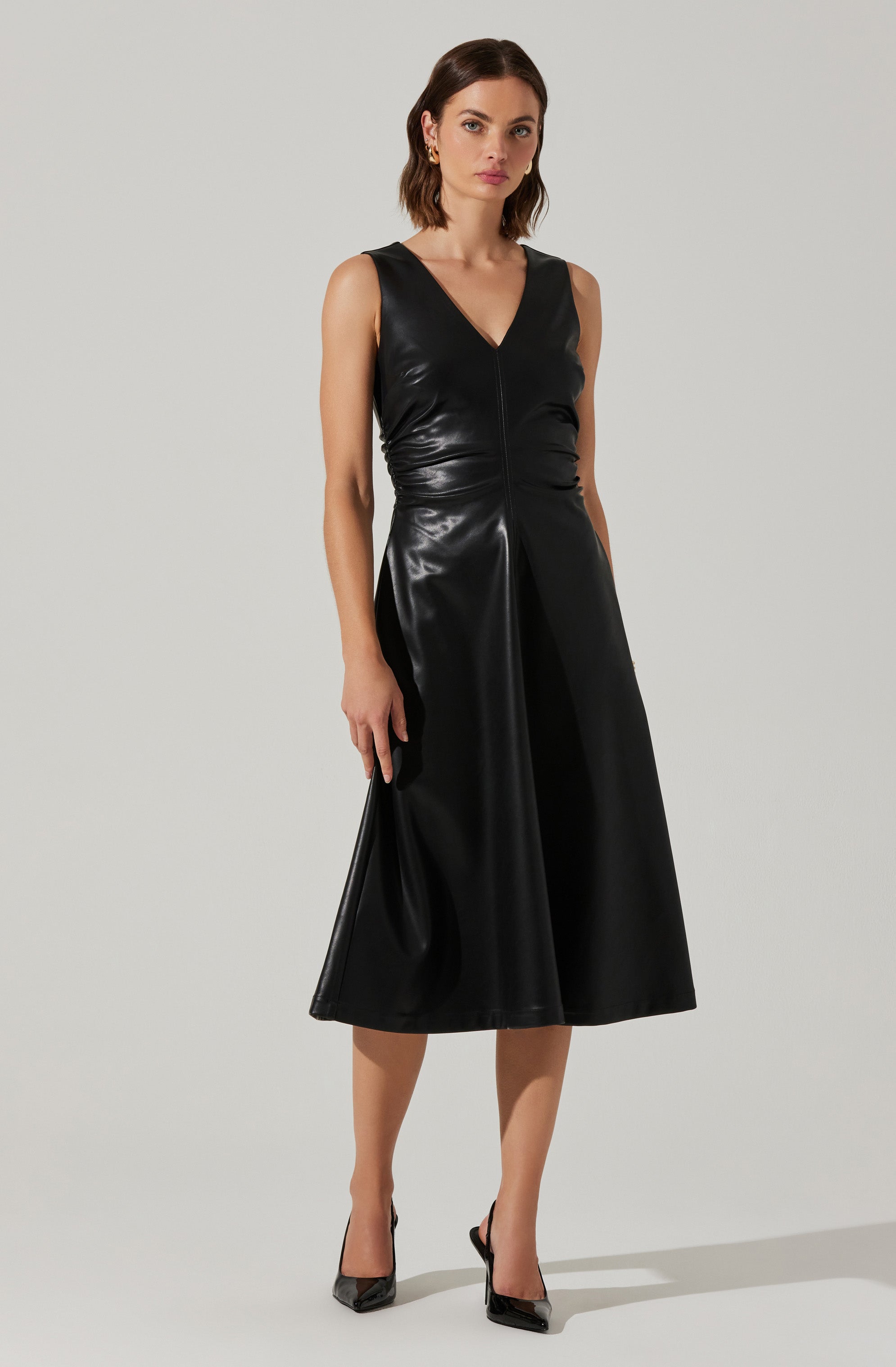 A woman is wearing a sleeveless black dress made of shiny material, featuring a V-neckline, a fitted bodice, and a high-waisted, A-line skirt that falls below the knees. She has on black pointed-toe high heels.