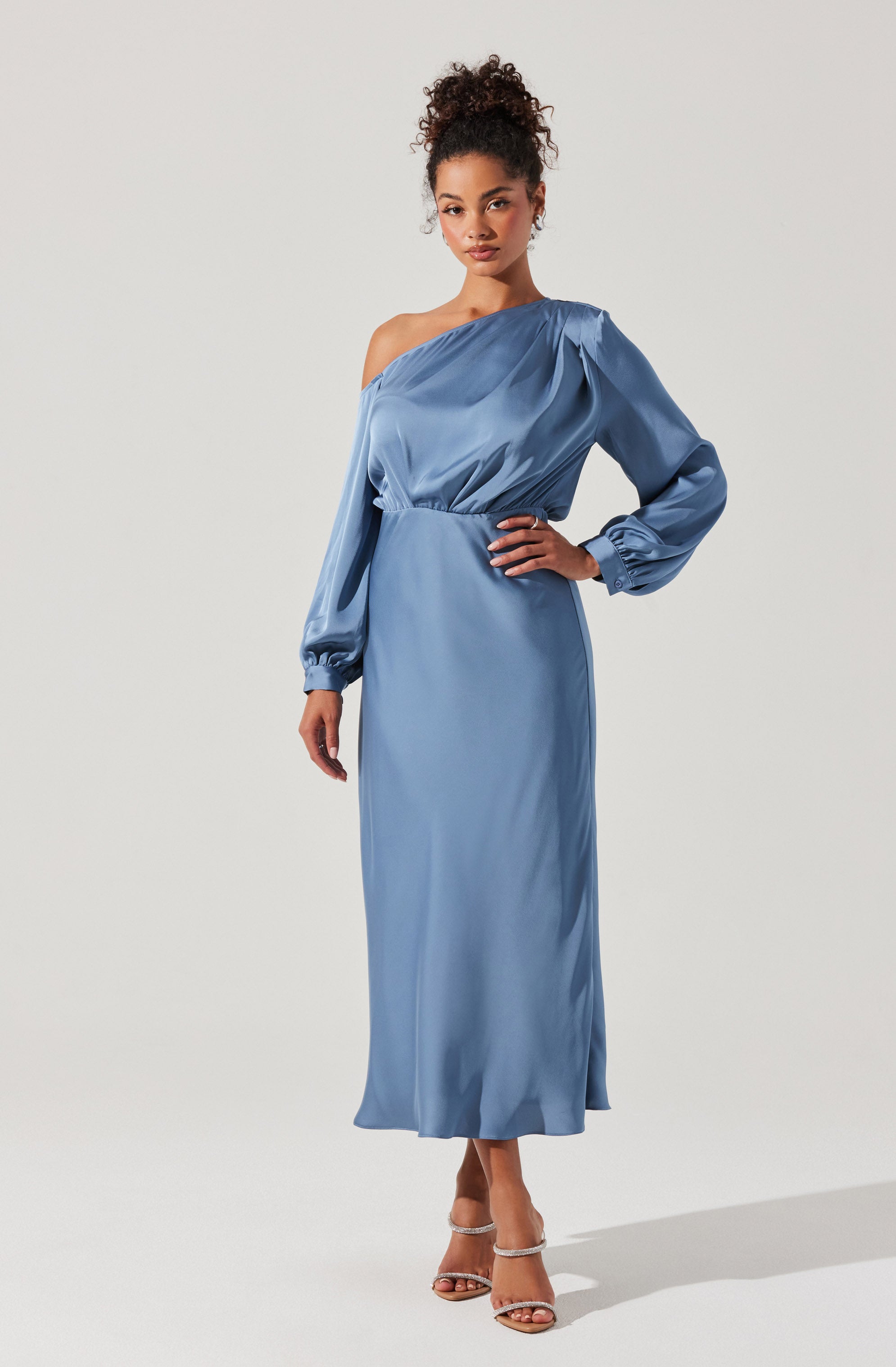 A woman is wearing a long, elegant, light blue satin dress. The dress features an asymmetrical neckline with one shoulder bare and the other covered by a long, puffed sleeve gathered at the wrist with elastic. The waistline is fitted, and the skirt flows down to her ankles. She is also wearing strappy high-heeled sandals.