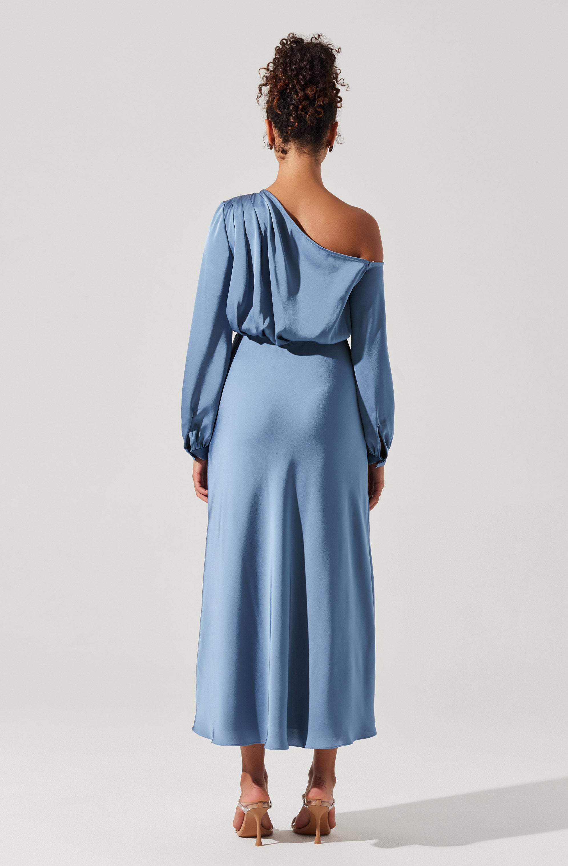 A woman is wearing a long, light blue satin dress with a one-shoulder design. The dress has long, slightly puffed sleeves with elasticized cuffs and a gathered detail on the shoulder. The skirt of the dress is flowing and reaches down to her ankles. She is wearing nude high-heeled shoes.