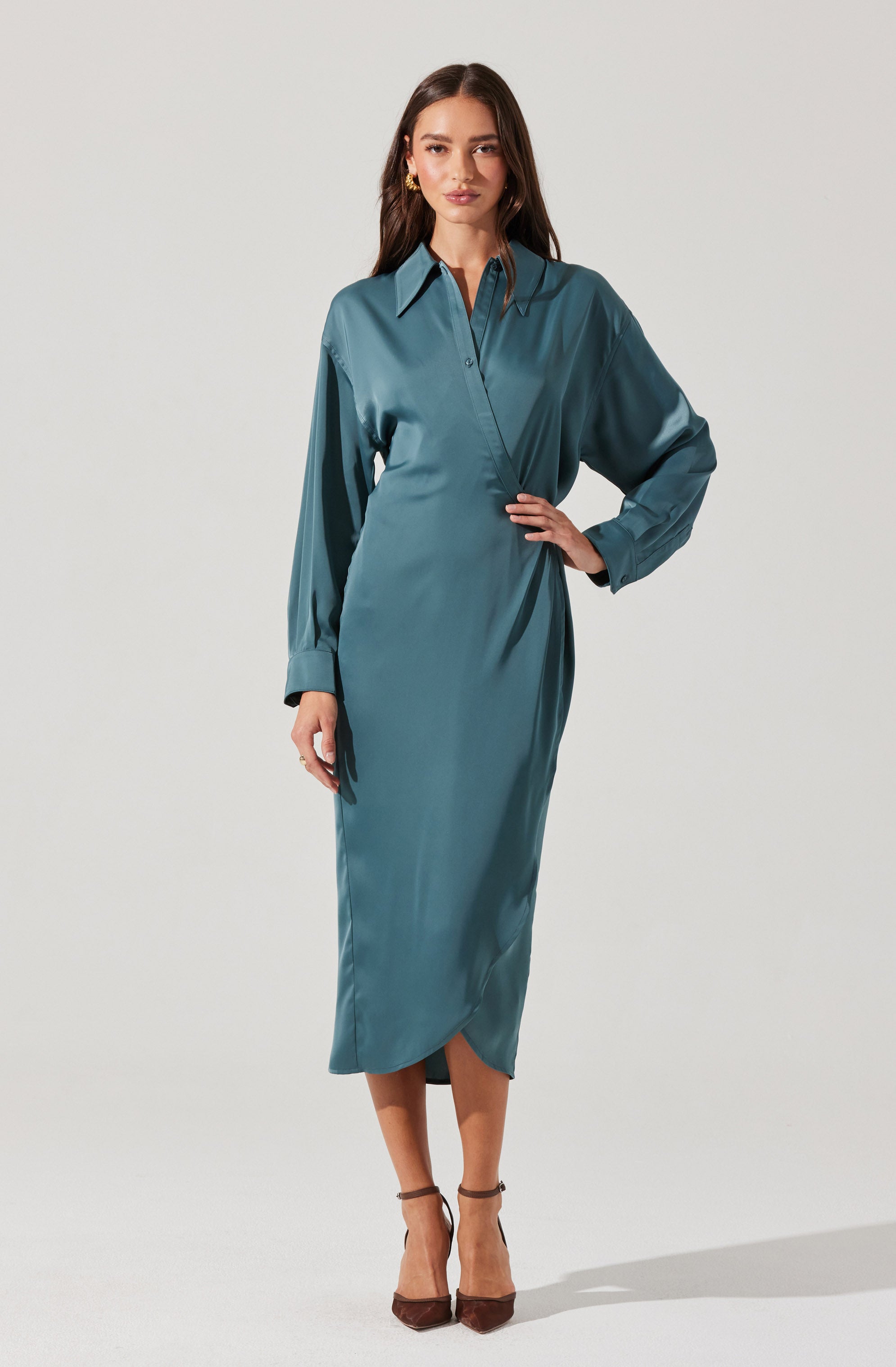 A woman is wearing a long, silky, teal-colored shirt dress with a pointed collar and button-down front. The dress has long sleeves with buttoned cuffs and a side slit at the hem. She is accessorizing with gold earrings and brown high-heeled shoes.