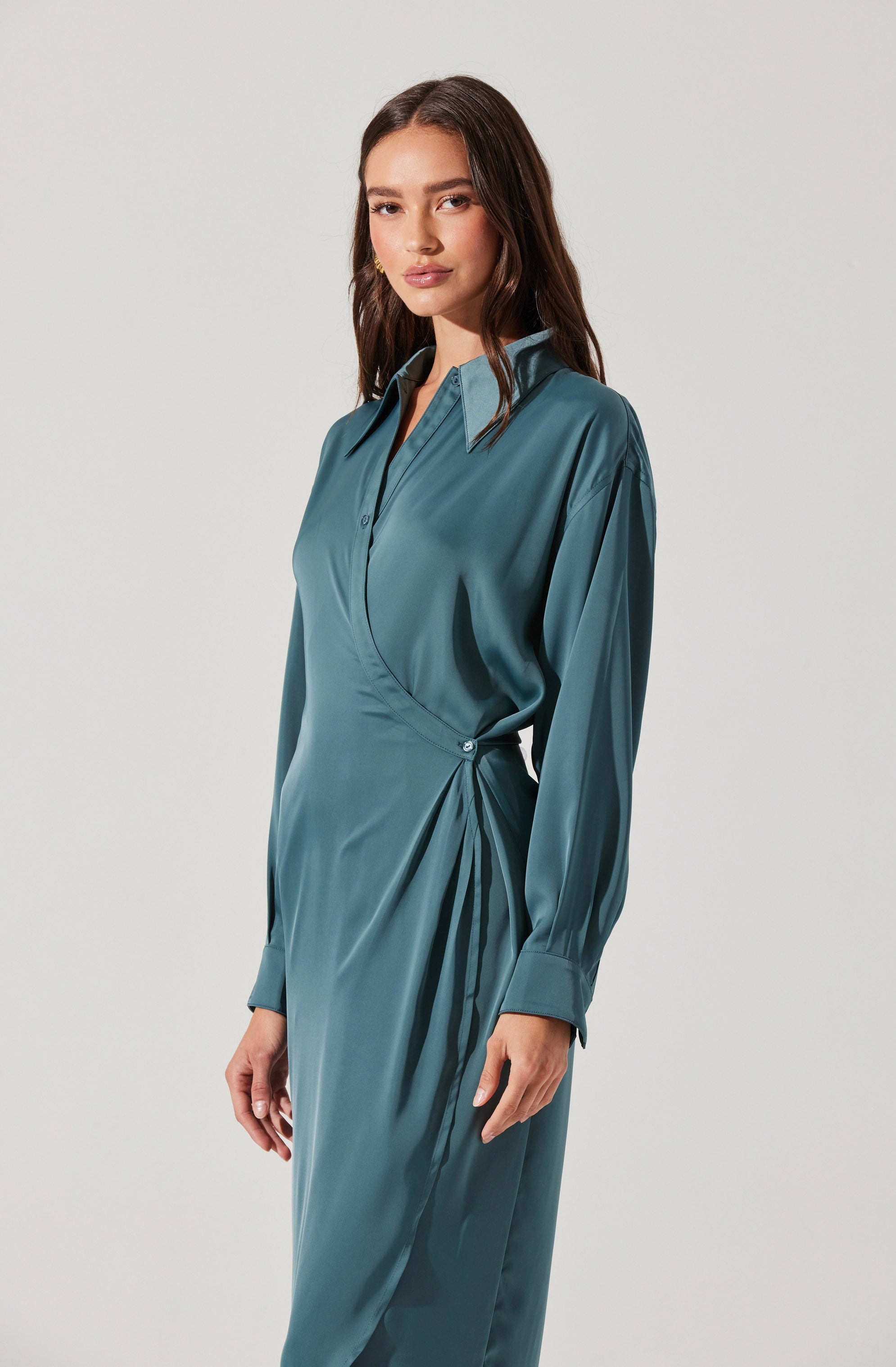 A woman is wearing a long, satin-like teal dress with a collar and buttons down the front. The dress has long sleeves with buttoned cuffs and features a draped, gathered detail at the waist, creating a wrap effect.