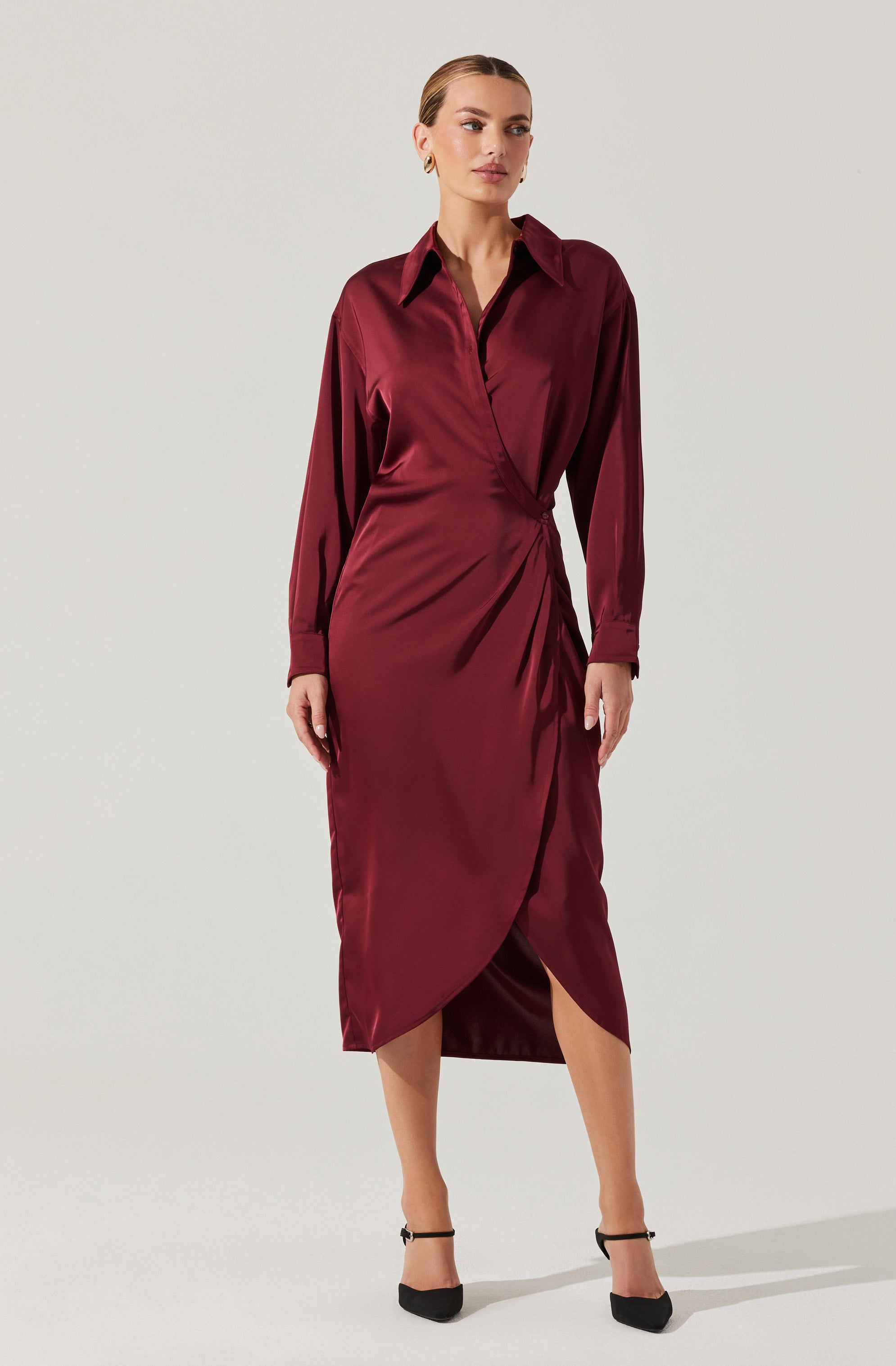 A woman is wearing a long-sleeved, deep burgundy satin dress with a classic collar and a wrap-style skirt that features an asymmetrical hemline with a slit. She is also wearing black high-heeled shoes with ankle straps.