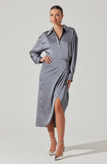 A woman is wearing a long, silver satin dress with a collared neckline and a button-down front. The dress has long sleeves that are slightly puffed at the shoulders and a high slit up one side, revealing her leg. She is also wearing high-heeled shoes with ankle straps.