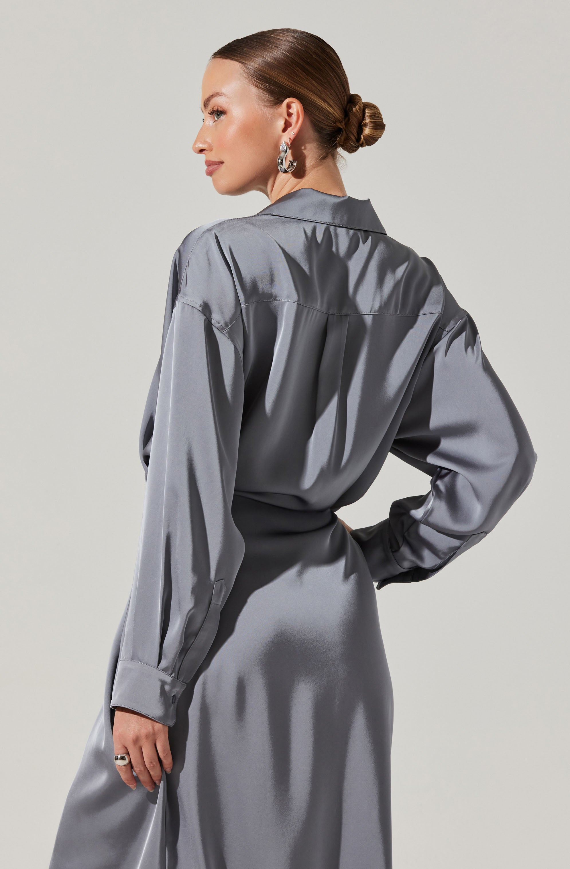 A woman is wearing a shiny, gray satin dress with long sleeves. The dress has a loose fit on the top with a collar, and the waist is cinched. She has her hair styled in a low, twisted bun and is accessorized with large, silver hoop earrings and a ring on her right hand.