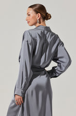 A woman is wearing a shiny, gray satin dress with long sleeves. The dress has a loose fit on the top with a collar, and the waist is cinched. She has her hair styled in a low, twisted bun and is accessorized with large, silver hoop earrings and a ring on her right hand.