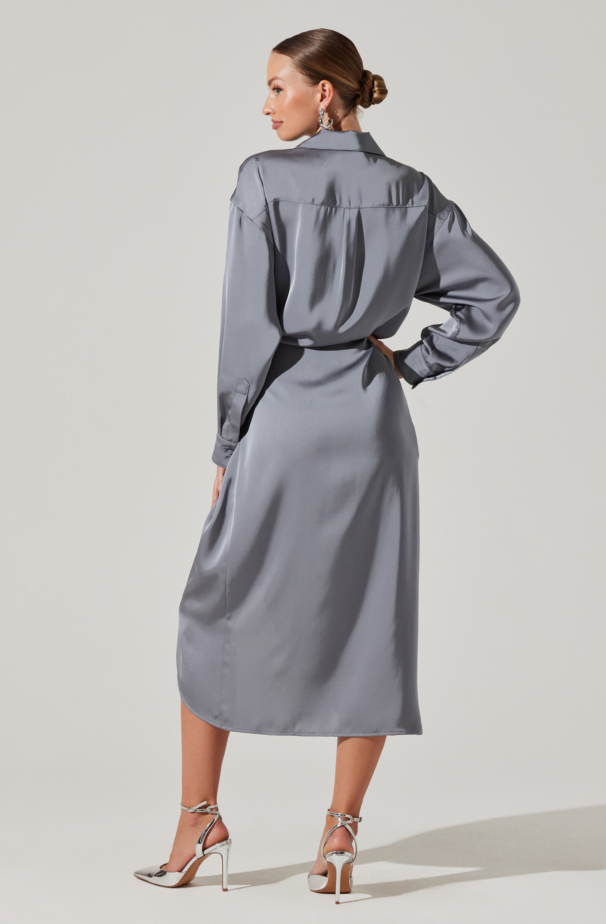 A woman is wearing an elegant, long, silver satin dress with a collared top and long sleeves that are gathered at the cuffs. The dress has a cinched waist and a flowing skirt that extends to her calves. She is also wearing high-heeled sandals with ankle straps, and her hair is styled in a sleek, low bun.