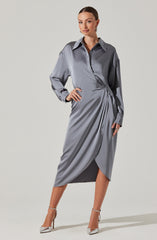 A woman is wearing a long, shiny, silver-gray satin dress with a collared neckline and long, slightly oversized sleeves. The dress features a draped design with pleats on one side, creating an asymmetrical, wrapped appearance. She is also wearing pointed silver high-heeled shoes with ankle straps.