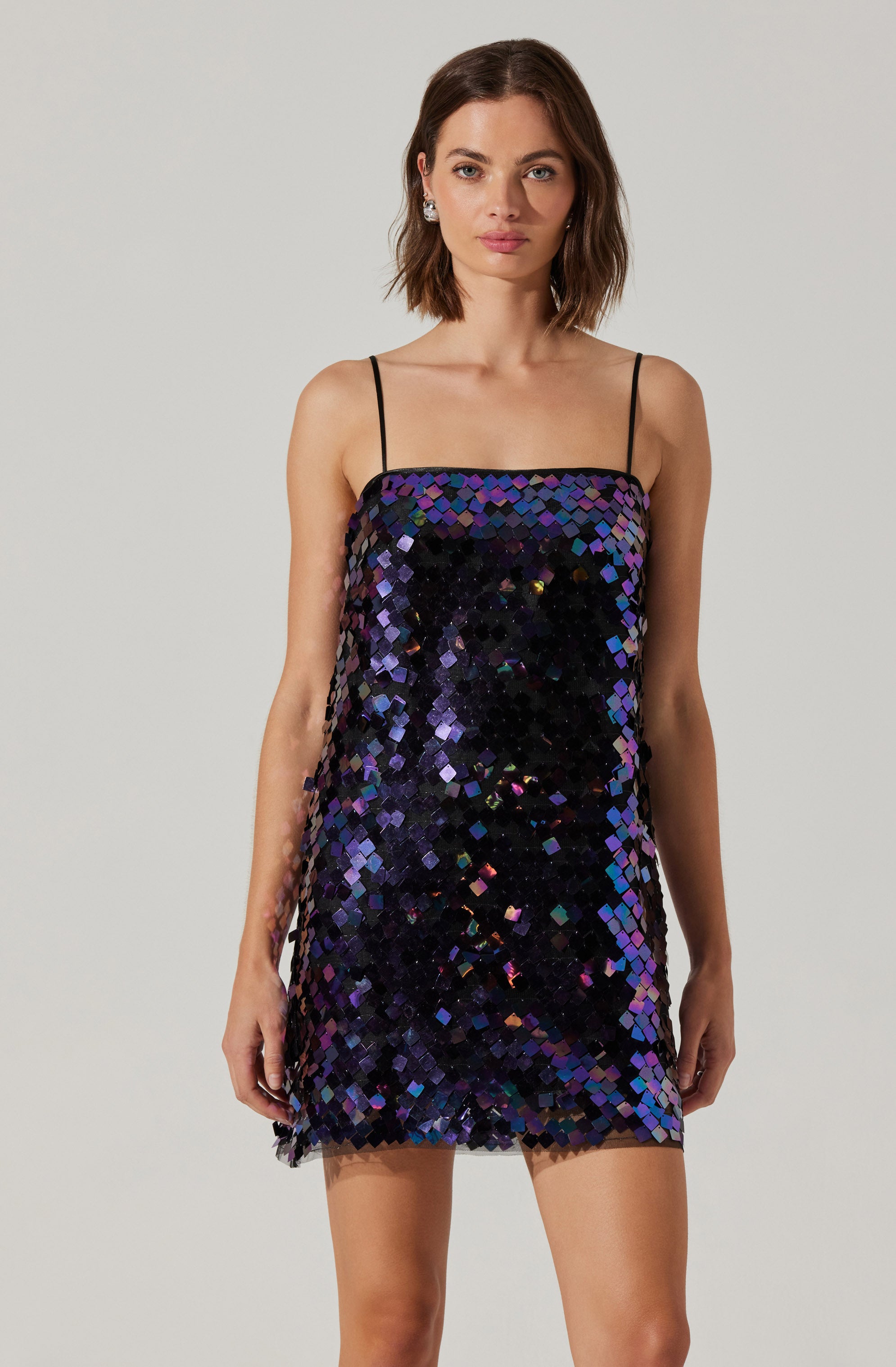 The dress is a sleeveless, mini-length garment with thin spaghetti straps. It features a straight neckline and is covered in large, iridescent sequins that shift colors between black, purple, and blue, creating a shimmering, reflective effect.