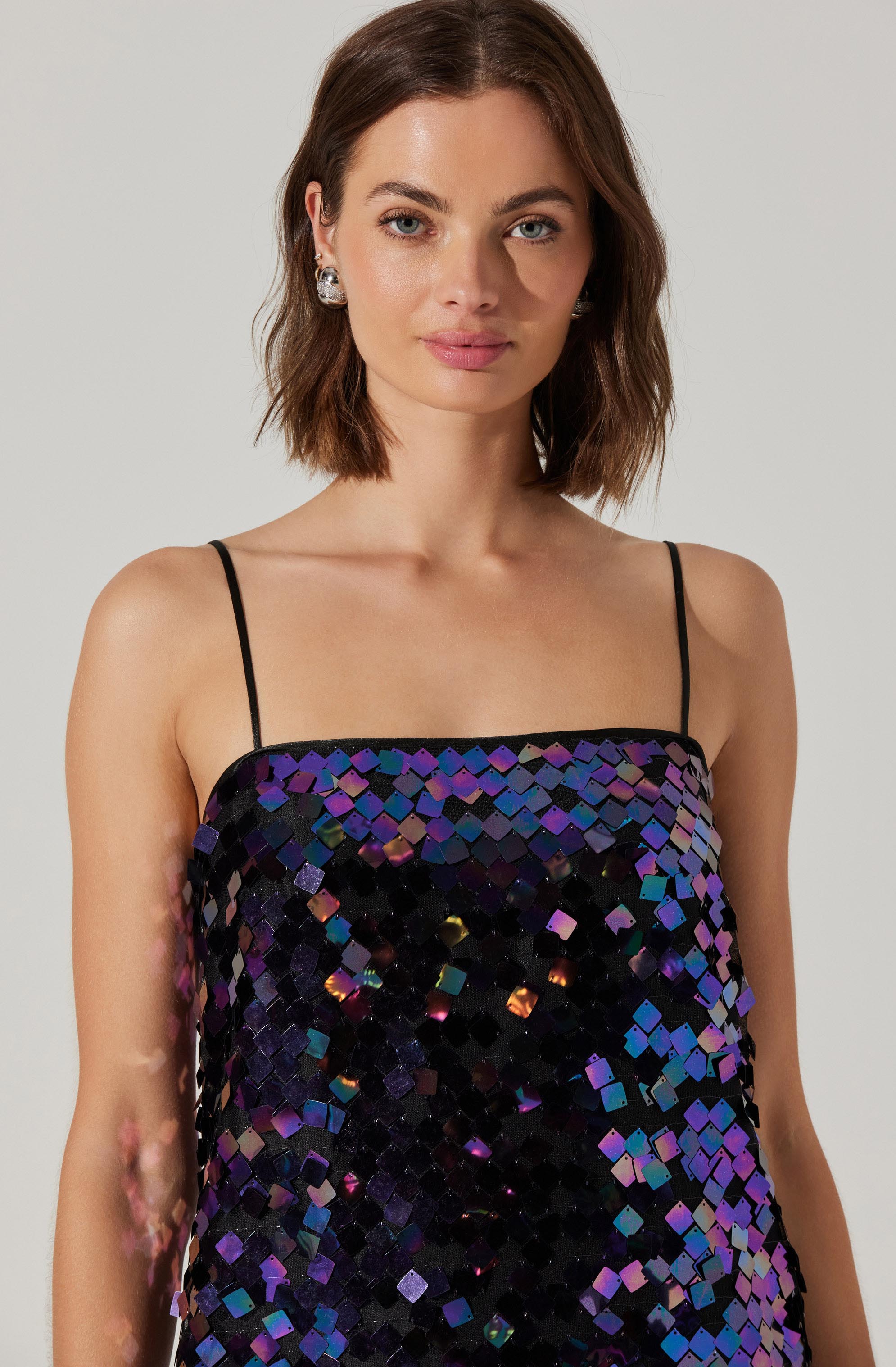 A woman with shoulder-length brown hair is wearing a strapless dress covered in large, multicolored sequins that reflect purple, blue, and hints of gold and pink. The dress has thin black spaghetti straps. She is also wearing large, silver, gemstone earrings.