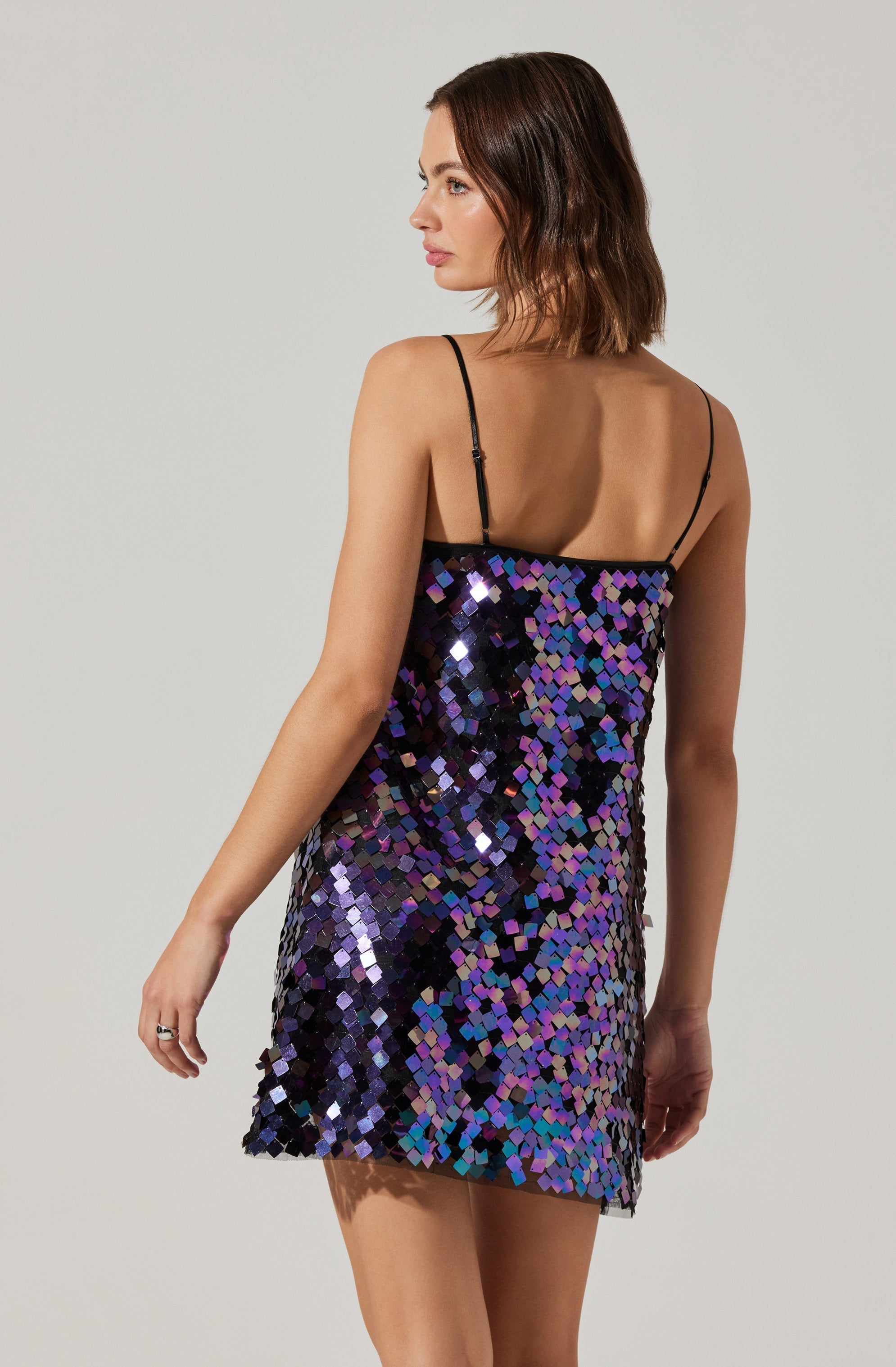 A woman is wearing a short, sleeveless dress with thin black spaghetti straps. The dress is covered in large, shiny, multicolored sequins in shades of purple, blue, and black, creating a sparkling, reflective appearance. The dress falls above the knees.