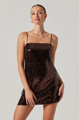 A woman is wearing a shiny, dark brown, sequined mini dress with thin spaghetti straps. The dress fits snugly and has a straight neckline. She is accessorized with gold hoop earrings and a ring on her right hand.