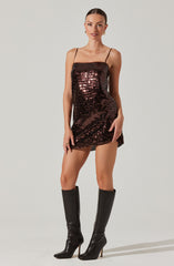 A woman is wearing a short, strapless dress covered in dark, shiny sequins that reflect light, creating a shimmering effect. The dress has thin shoulder straps and a fitted silhouette. She pairs the dress with tall, black, high-heeled boots that reach just below her knees.
