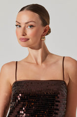 A woman is wearing a sleeveless, square-neck dress with thin spaghetti straps. The top part of the dress is covered in dark, shiny sequins, giving it a textured and reflective appearance. She has gold hoop earrings and a neat, low bun hairstyle.