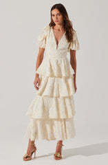 A woman is wearing an elegant, cream-colored dress made of lace fabric. The dress features a deep V neckline with scalloped edges, short puffed sleeves, and a fitted waist. The skirt is tiered with multiple layers of ruffled lace, creating a flowing, layered look that extends to her ankles. She is also wearing high-heeled sandals with open toes and floral embellishments.
