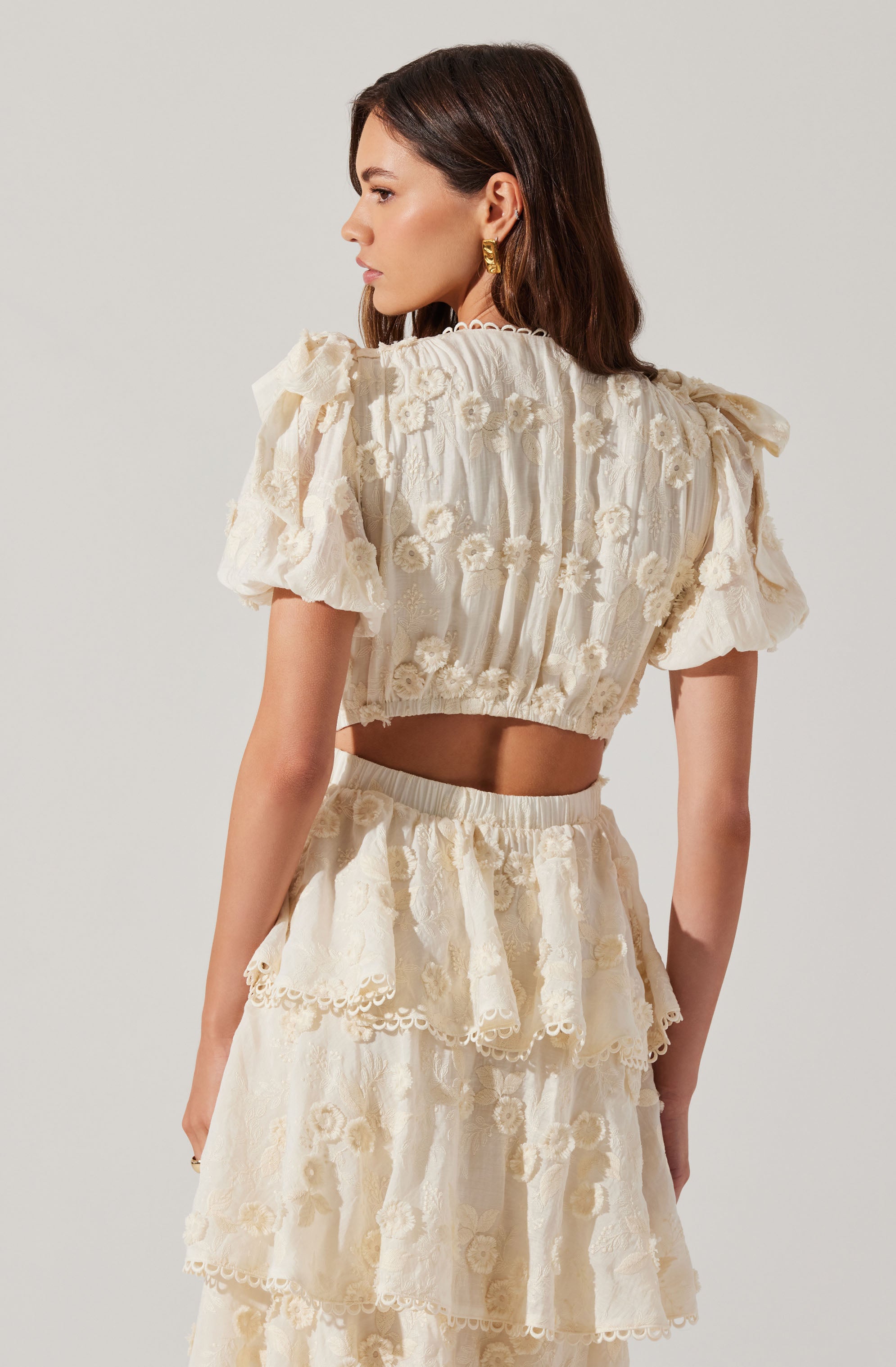 A woman is wearing a matching cream-colored two-piece outfit consisting of a cropped top with puffed short sleeves and a high-waisted skirt. Both pieces feature intricate floral embroidery and lace detailing. The top has a loose fit with ruffled accents, and the skirt includes layered, ruffled tiers with decorative edges.