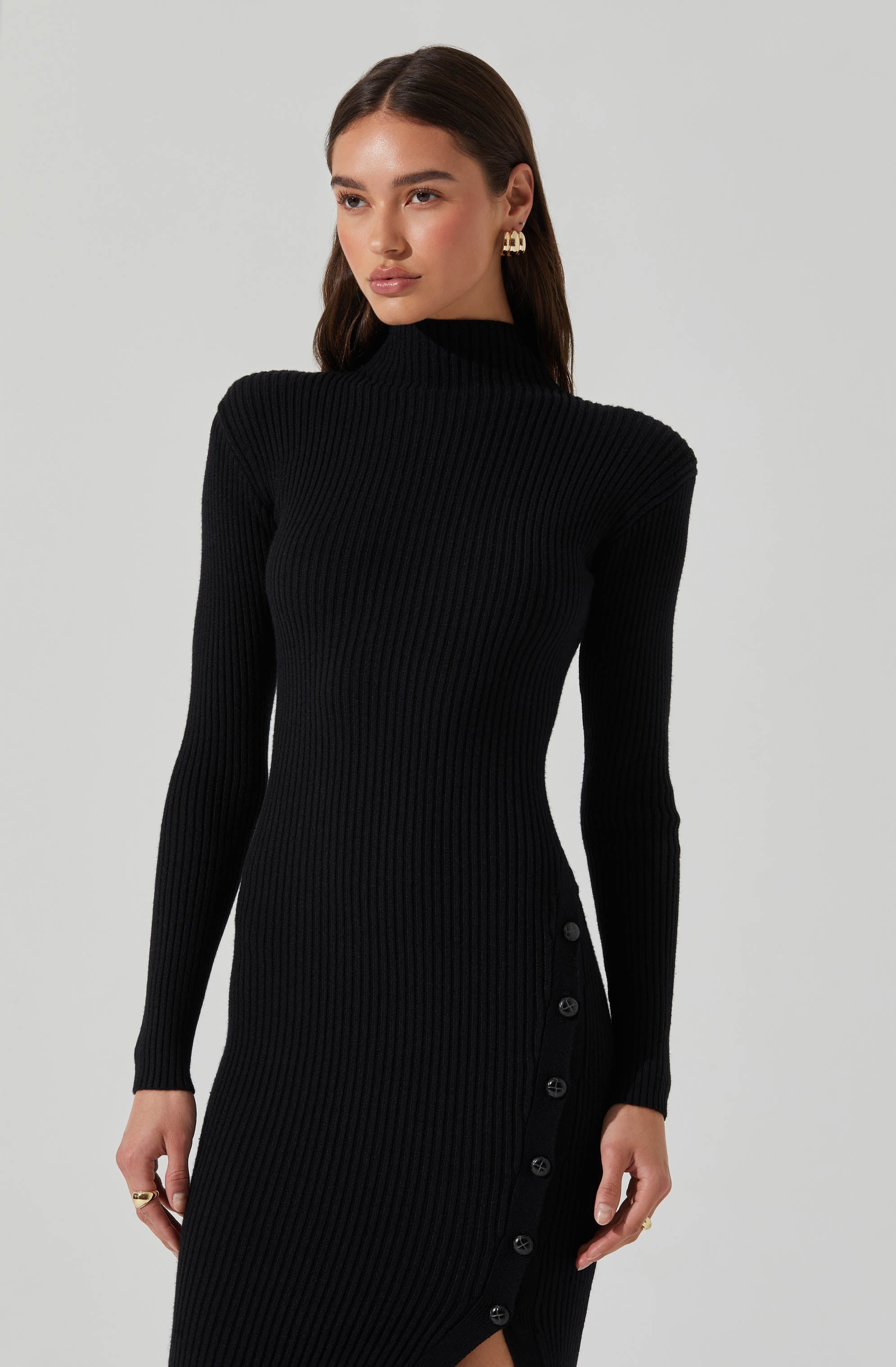 A woman is wearing a fitted, long-sleeve, black ribbed knit dress with a high neckline. The dress features a row of black buttons along the side, extending from the waist to the hem, creating a slit. She is accessorized with gold earrings and rings.