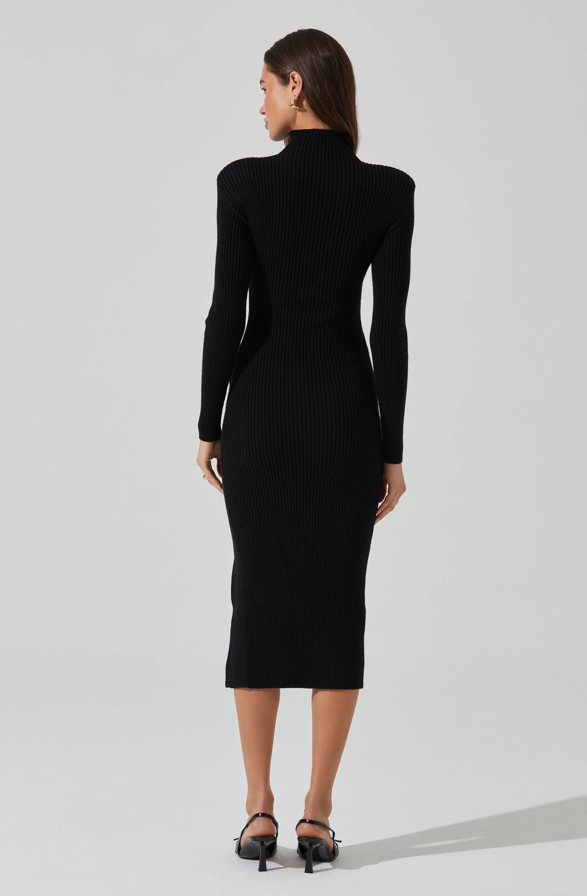 A woman is wearing a fitted, long-sleeve, ribbed black dress that extends to mid-calf. The dress features a high neckline and a sleek, body-hugging style. She is also wearing black high-heeled shoes with ankle straps.