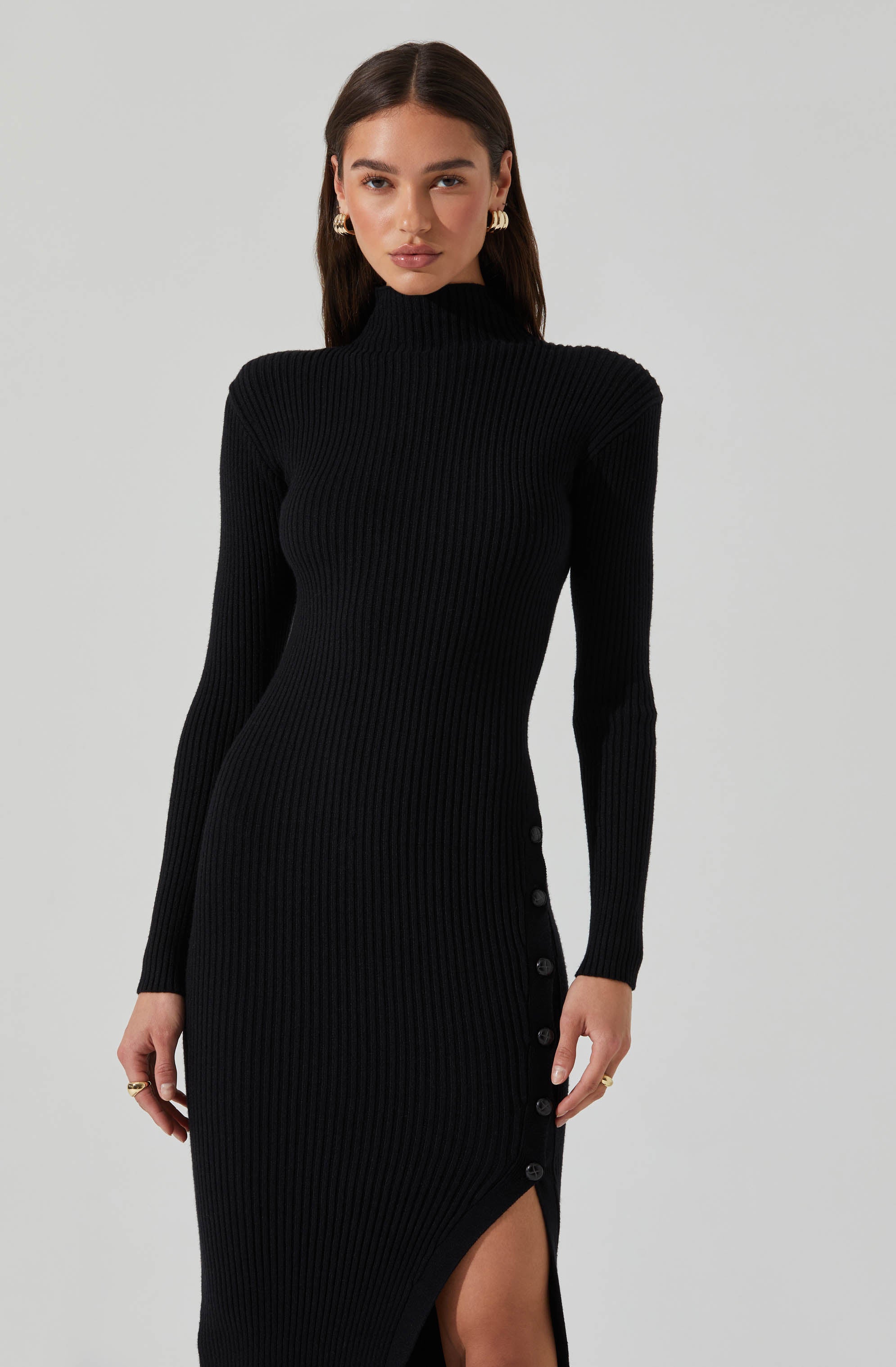A woman is wearing a fitted, long-sleeved, ribbed black dress with a high turtleneck. The dress features buttons along the side from the waist down to the hem, where there is a slit that reveals part of her thigh.