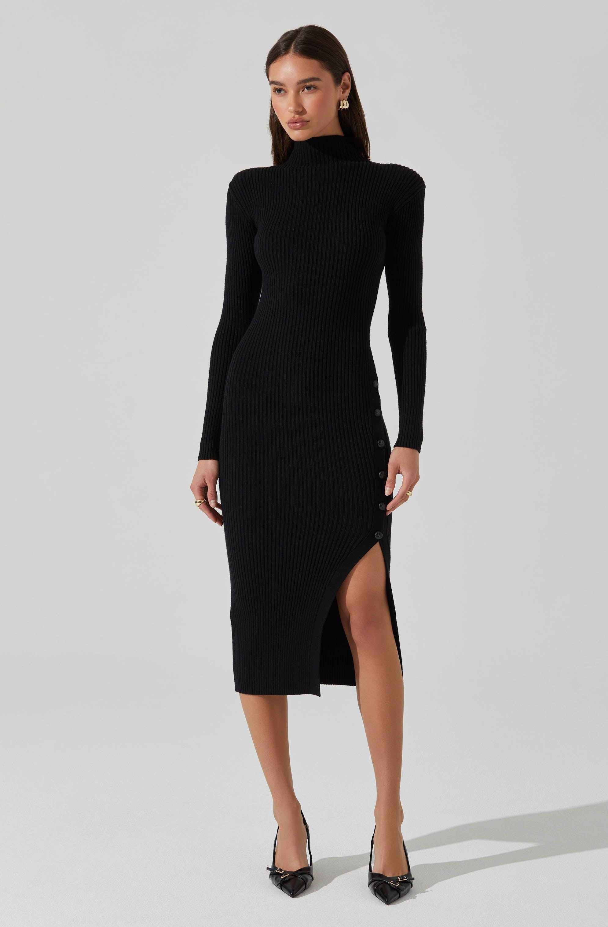 A woman is wearing a black, long-sleeved, ribbed knit dress with a high neckline. The dress features a side slit with buttons along the opening. She is also wearing black pointed-toe high heels with ankle straps and gold hoop earrings.
