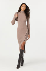 A woman is wearing a beige, long-sleeved, ribbed knit dress that reaches mid-calf. The dress features a high neckline, a side slit with large buttons running alongside it, and a fitted silhouette. She has one shoulder slightly raised with her hand holding a strap of a small brown shoulder bag. She is also wearing tall black leather boots that reach above her knees.