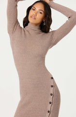 A woman is wearing a fitted, ribbed knit dress in a taupe color, featuring a high turtleneck and long sleeves. The dress has a row of dark buttons along the side from the waist to the hem.