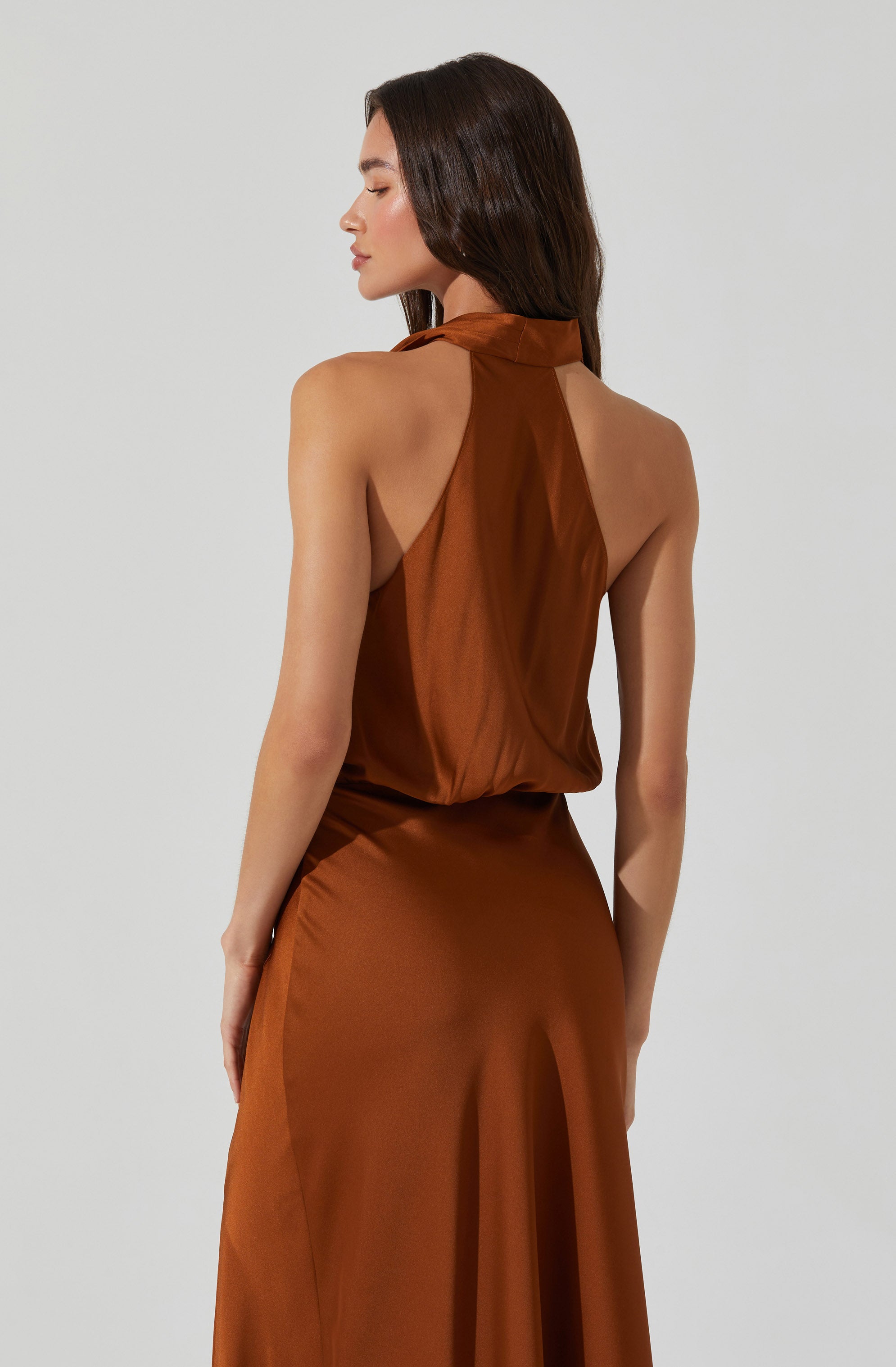 A woman is wearing a sleeveless, high-neck, satin dress in a warm, bronze-brown color. The dress has a draped, loose-fitting top and a fitted waist. The skirt is long and flowing, with a smooth, shiny surface that reflects light.