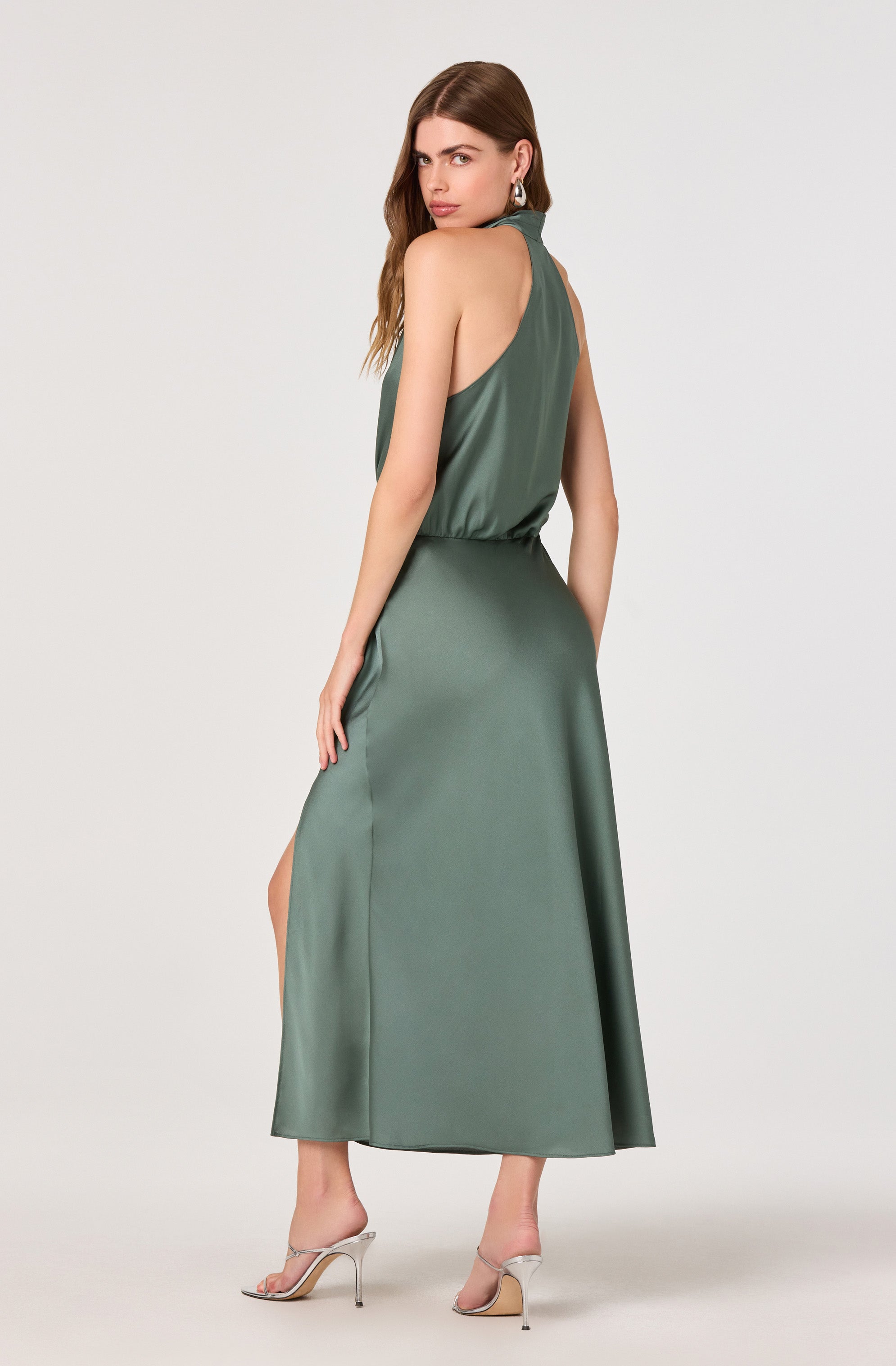 A woman is wearing a sleeveless, high-neck, satin dress in a muted green color. The dress has a loose-fitting bodice with a gathered waist and a long, flowing skirt that reaches the floor. The back of the dress features a keyhole opening at the neckline. She is also wearing a pair of clear high-heeled sandals.