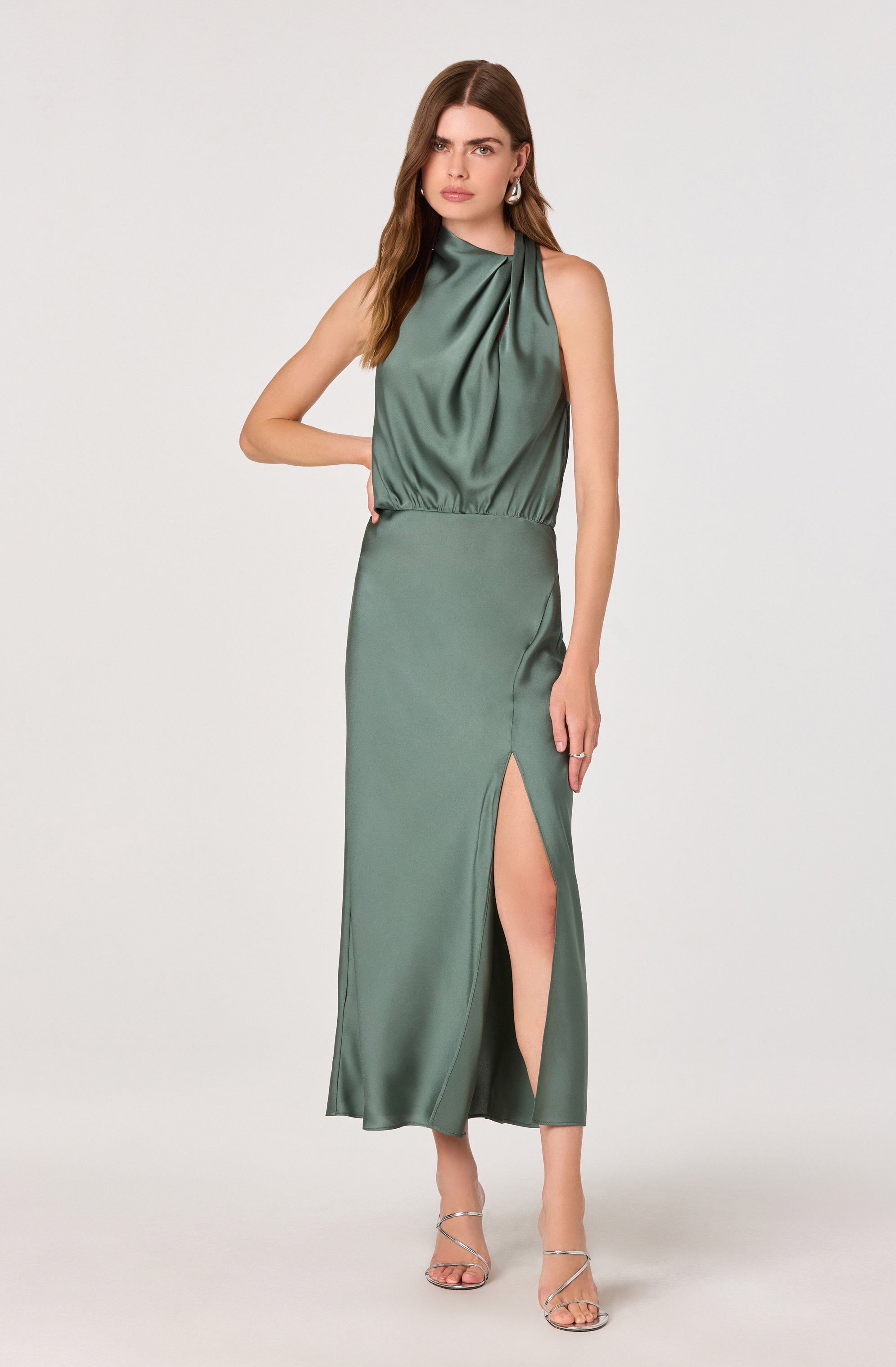 A woman wearing a sleeveless, satin, green dress with a high neckline featuring gathered fabric at the top. The dress has a fitted waist and a long skirt with a high slit on the left side that reaches mid-thigh. She is also wearing strappy high-heeled sandals.