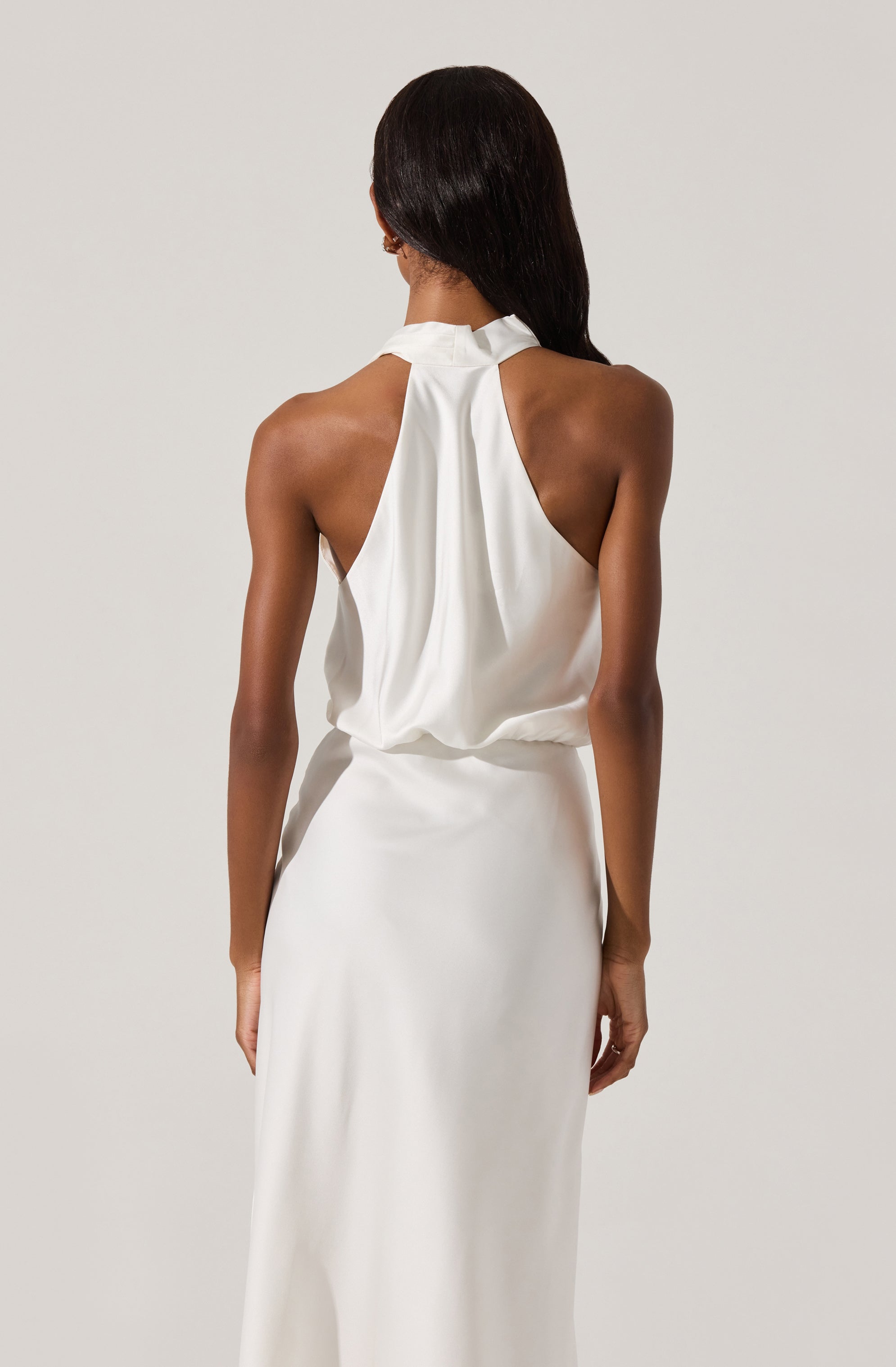 A woman is wearing a sleeveless, high-neck, white satin dress with a loose draped top and a fitted, floor-length skirt. The back of the dress features a gathered, flowing design.