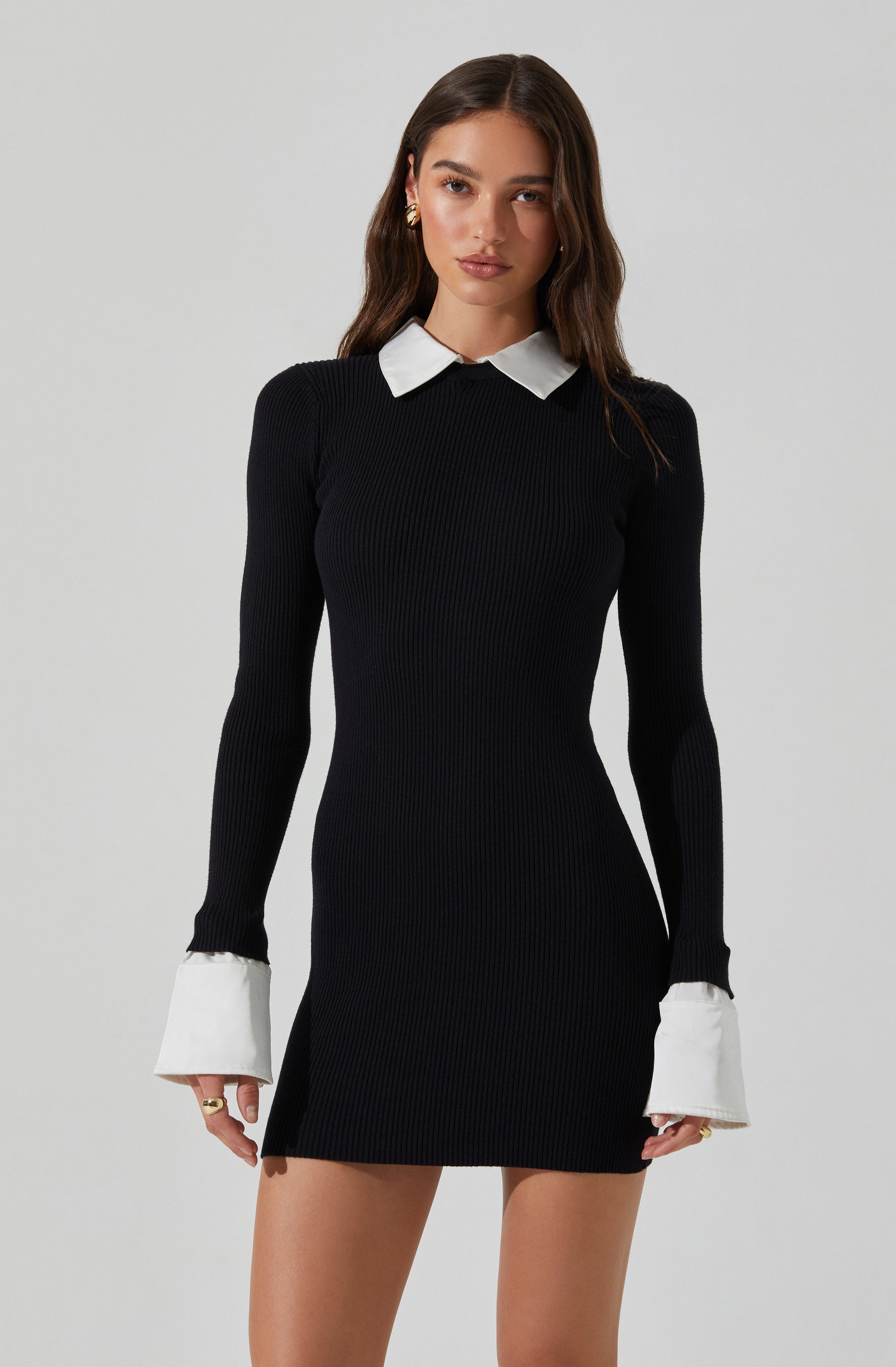 A woman is wearing a fitted, long-sleeve black ribbed knit dress with white contrasting collar and cuffs. The dress is short, ending above the knees, and the cuffs are designed with added white fabric that extends beyond the sleeves.