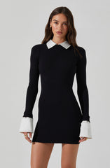 A woman is wearing a fitted, long-sleeve black ribbed knit dress with white contrasting collar and cuffs. The dress is short, ending above the knees, and the cuffs are designed with added white fabric that extends beyond the sleeves.