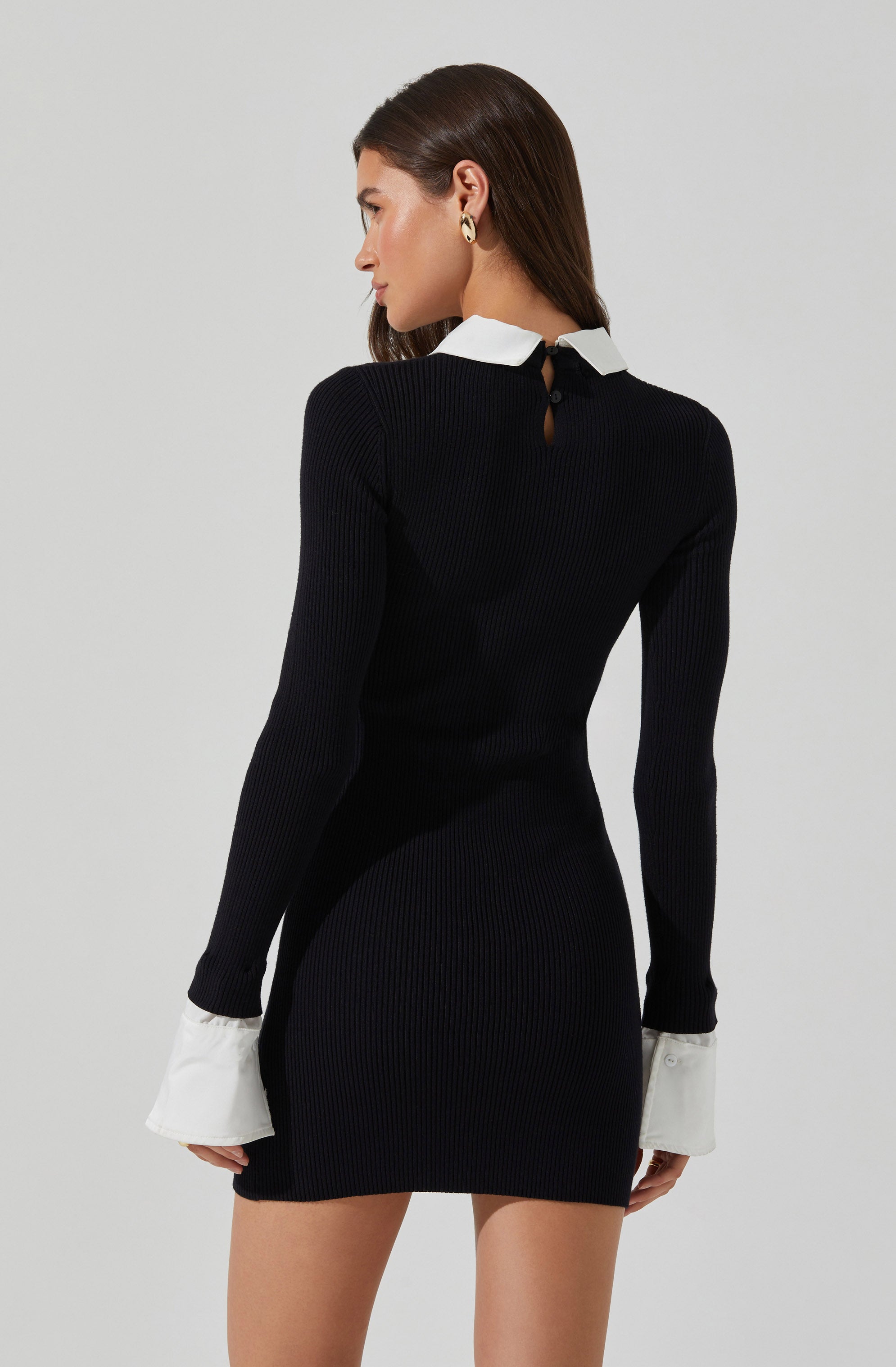 A woman is wearing a fitted, long-sleeved black ribbed knit dress with white cuffs at the sleeve ends and a contrasting white collar. The dress features a small keyhole opening at the back, fastened with a button.